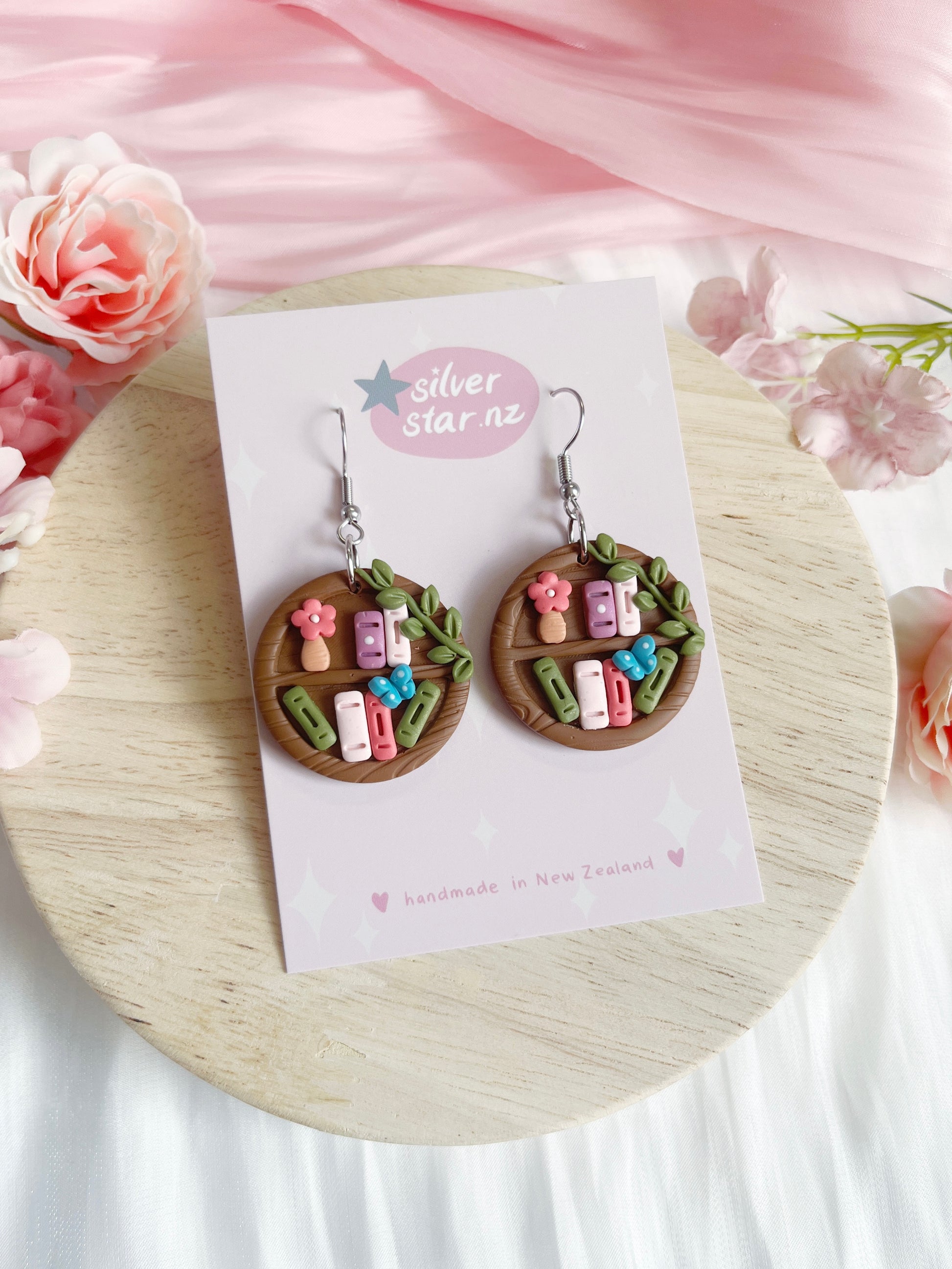 Round Bookshelf Polymer Clay Earrings by silverstar.nz: handmade, featuring mini pastel books, flowers, and leaves on brown circular bases. Displayed on a branded card with pink floral accents and a light wood background.