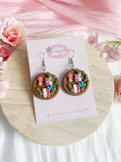 Round Bookshelf Polymer Clay Earrings by silverstar.nz: handmade, featuring mini pastel books, flowers, and leaves on brown circular bases. Displayed on a branded card with pink floral accents and a light wood background.