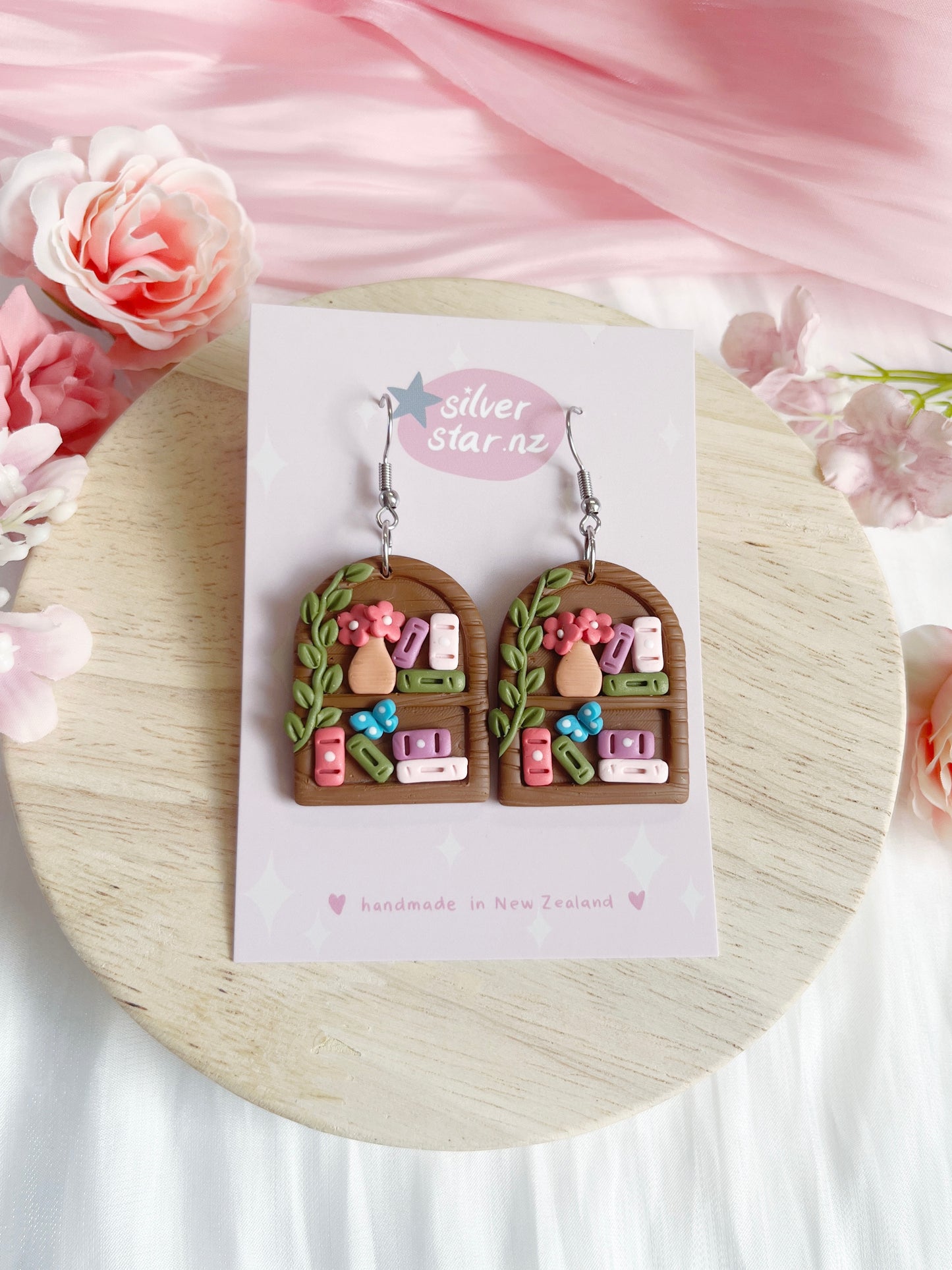 The Round Arch Bookshelf Polymer Clay Earrings by silverstar.nz feature brown arched bookshelf shapes with colorful mini books, plant vine, flowers, vase details, stainless steel hooks, and come on a branded pink-floral backing card.