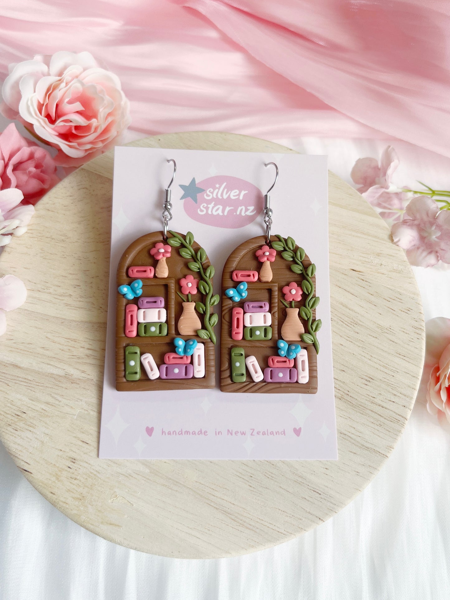 Tall Arch Bookshelf Polymer Clay Earrings by silverstar.nz feature a funky arched door design with colorful books, flowers, and leaves. Presented on a branded card with pink flowers and a light wood background.
