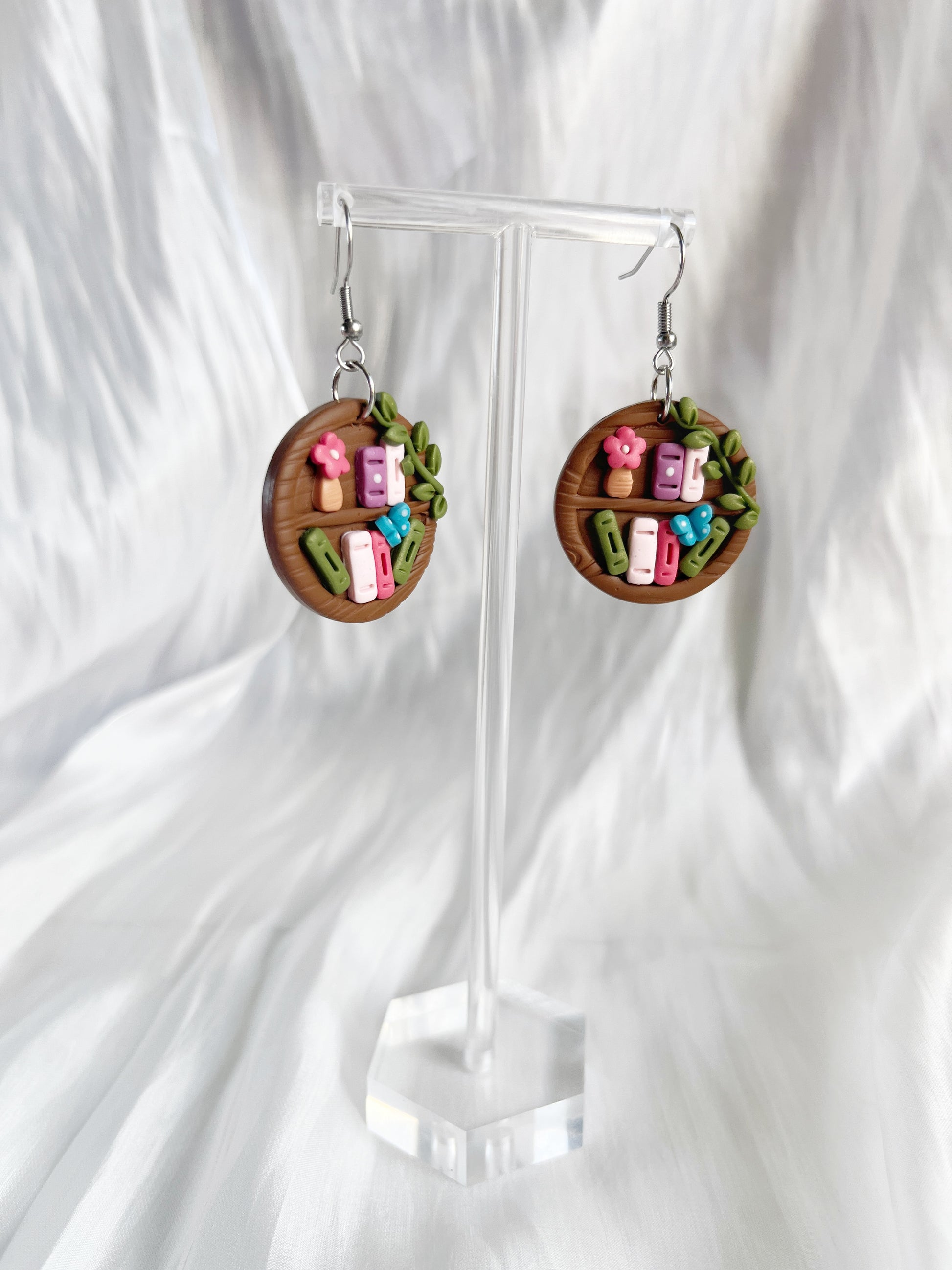 Round Bookshelf Polymer Clay Earrings by silverstar.nz are handmade, featuring miniature books, flowers, and leaves on a wooden-look base. Displayed on a clear stand against a soft white textured fabric.