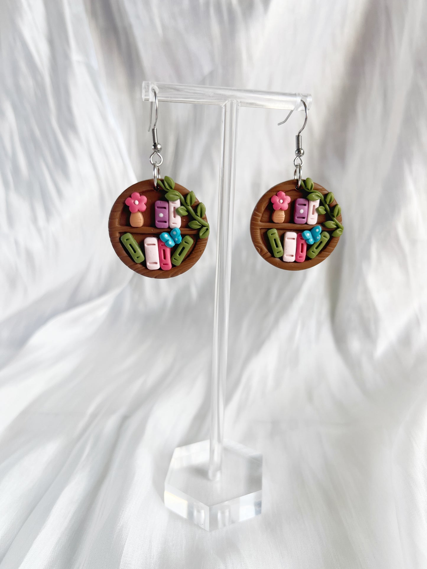 Round Bookshelf Polymer Clay Earrings by silverstar.nz showcase miniature sushi, leaves, and pink flowers on a wood-look background, displayed on a clear stand with a white fabric backdrop.