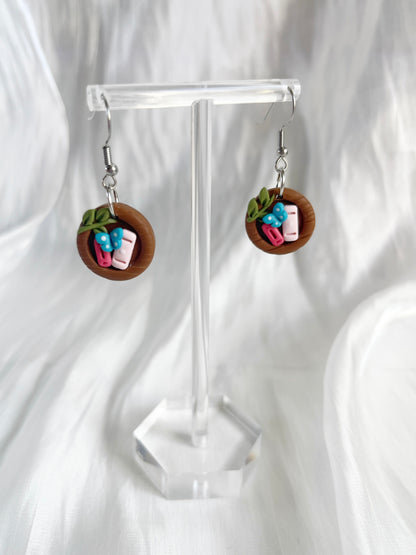 The Mini Round Bookshelf Polymer Clay Earrings by silverstar.nz feature a unique wooden plate design with miniature sushi rolls, including pink fish and green leaves, displayed on a transparent stand against a white background.