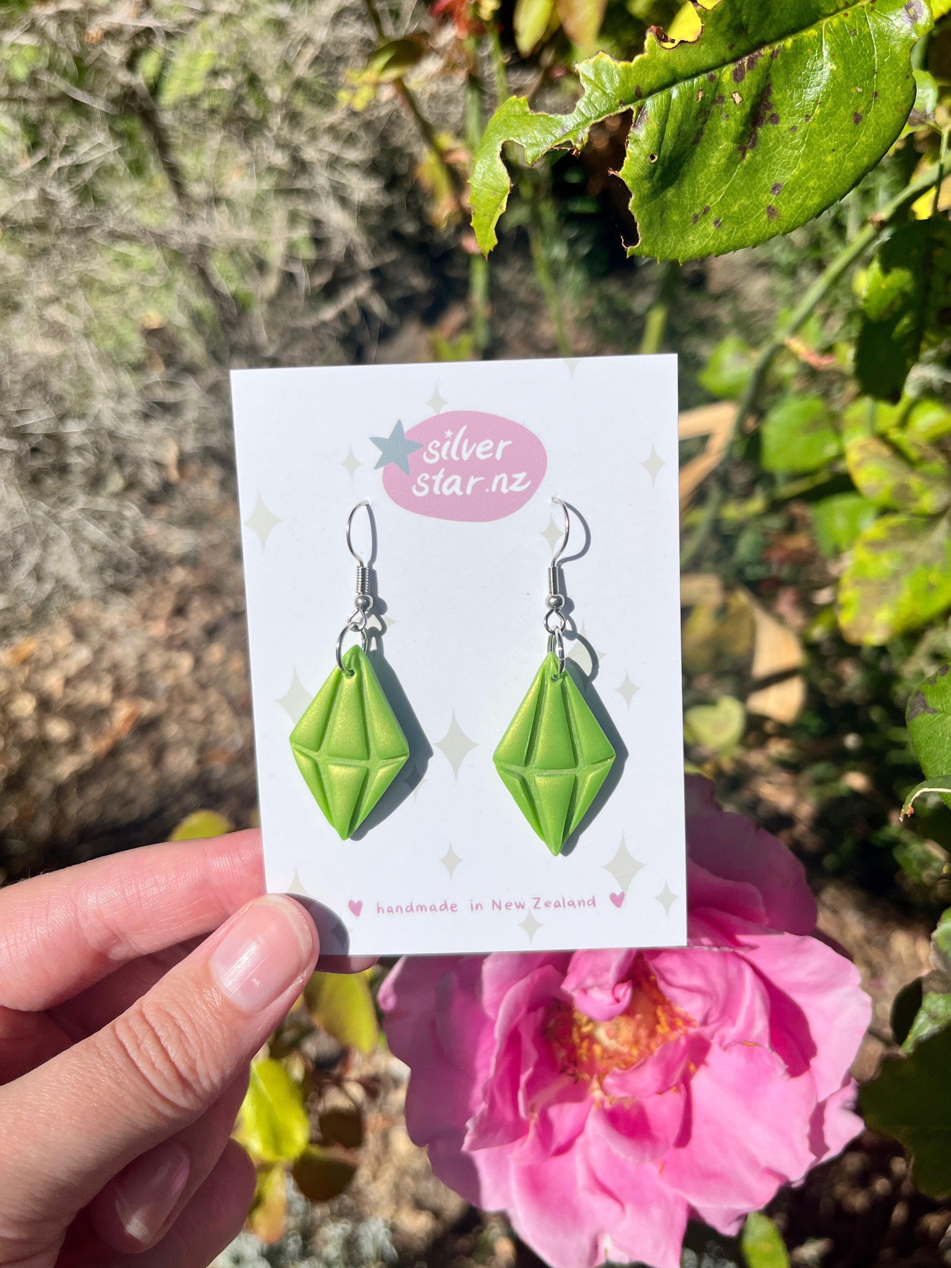 A hand holds a card with Sims Plumbob Polymer Clay Earrings by silverstar.nz in front of a blooming pink rose and green foliage. The card reads silverstar.nz and handmade in New Zealand.