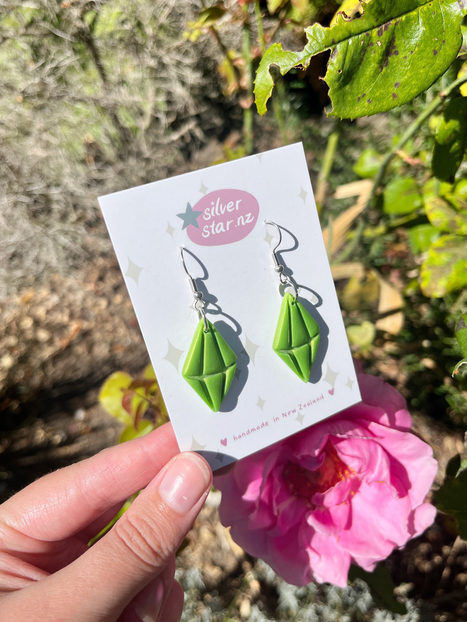 A hand holds a card featuring Sims Plumbob Polymer Clay Earrings from silverstar.nz in front of a pink rose and green leaves in sunlight; the card notes theyre handmade in New Zealand.