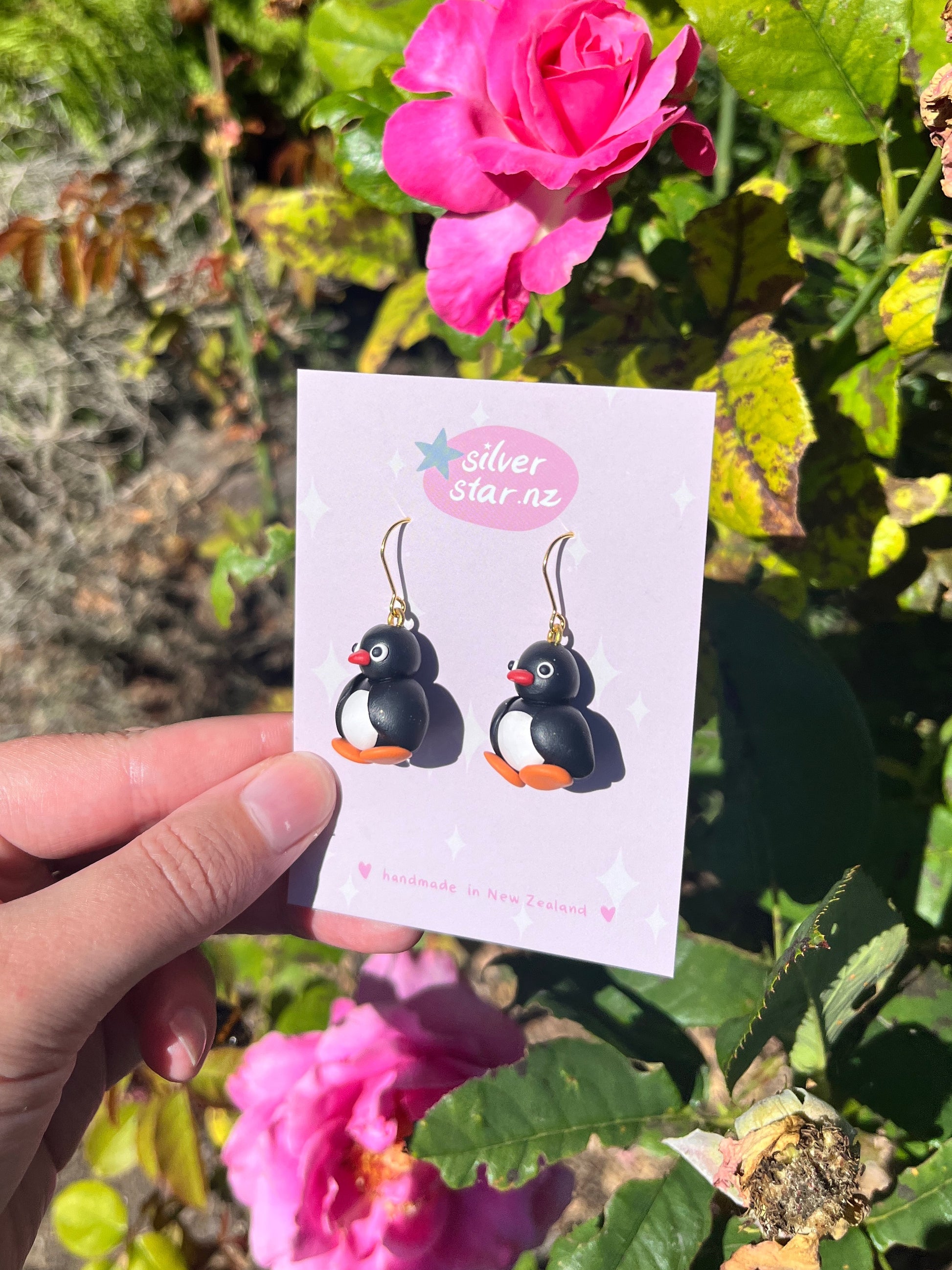 A hand displays Pingu Polymer Clay Earrings amid vivid pink flowers and green leaves. The card reads silverstar.nz and states handmade in New Zealand, emphasizing the quirky, funky design.
