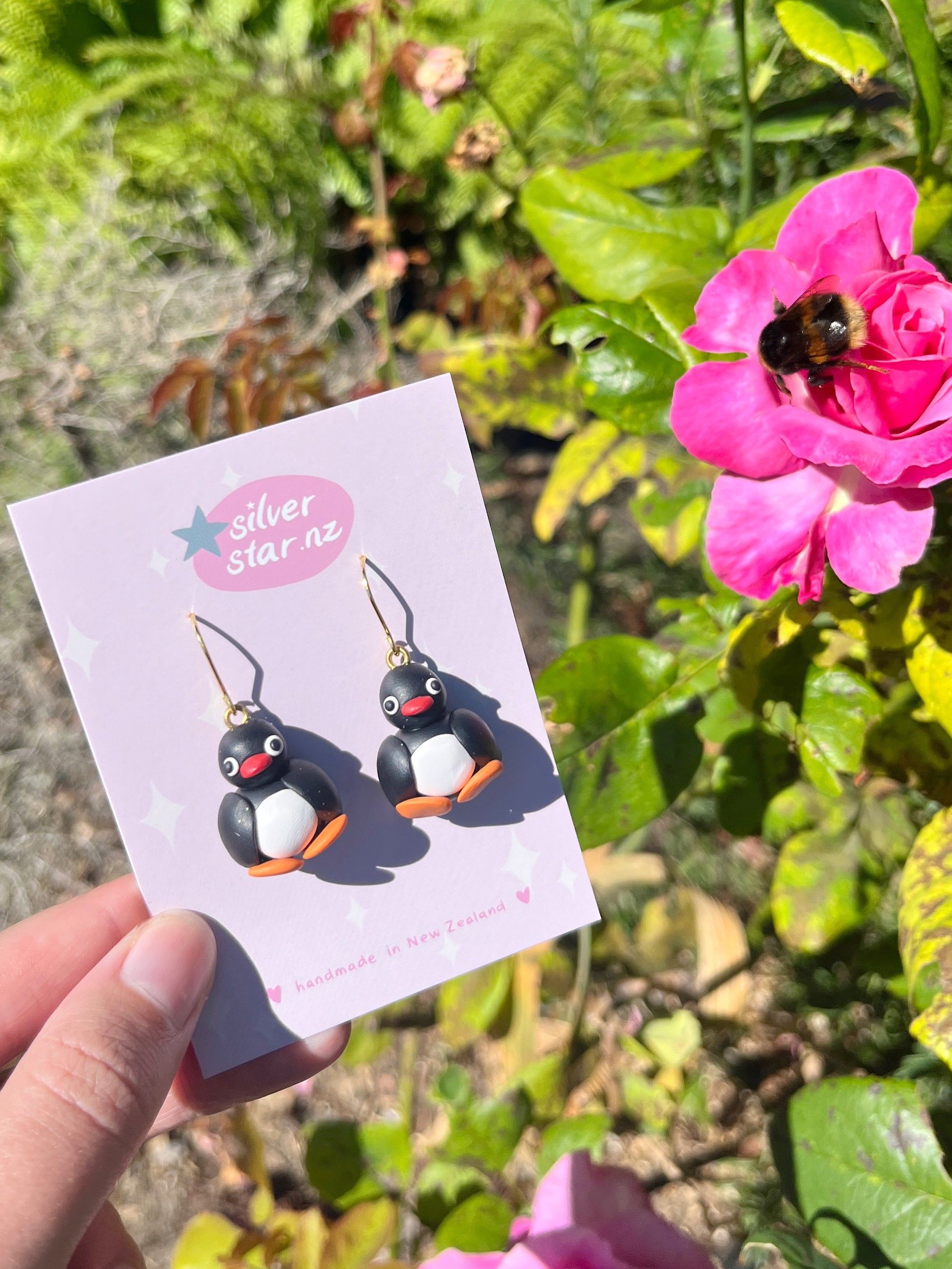 A hand holds Pingu Polymer Clay Earrings by silverstar.nz against a card, surrounded by a garden with a bee hovering near a vibrant pink flower.