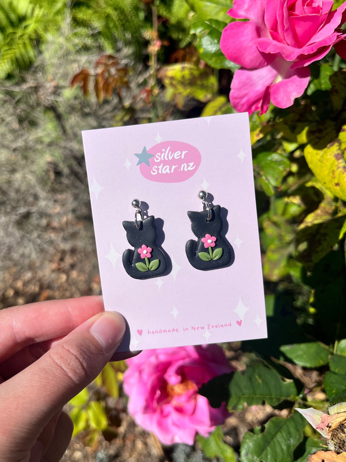 A hand holds a silverstar.nz card with Black Cat Polymer Clay Earrings featuring pink flowers, near vibrant pink roses and green leaves in sunlight. The card reads silverstar.nz handmade in New Zealand.