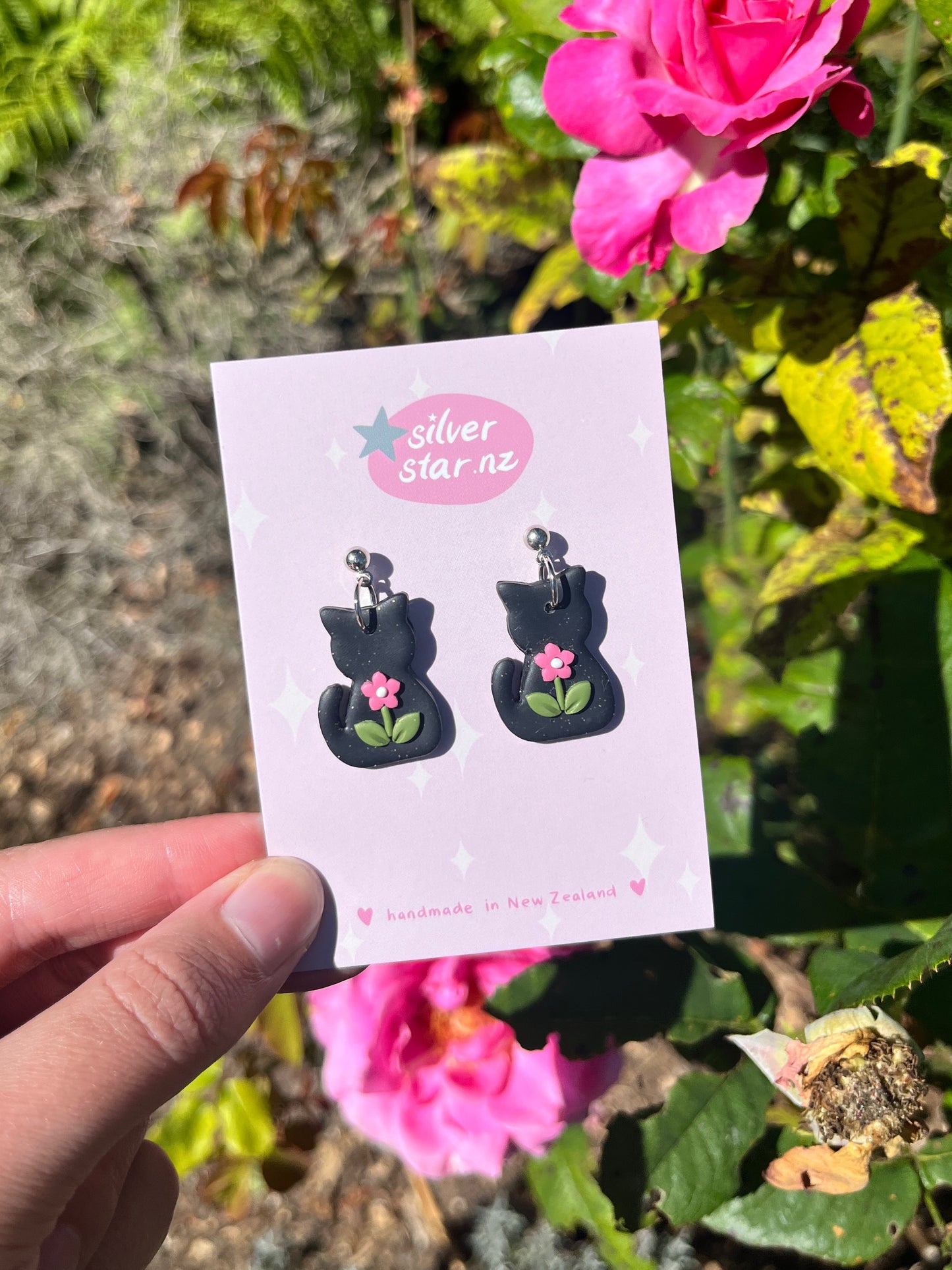 A hand holds a card showing Black Cat Polymer Clay Earrings with pink flowers from silverstar.nz. Pink roses and green leaves are in the sunny background. The card says silverstar.nz and handmade in New Zealand.