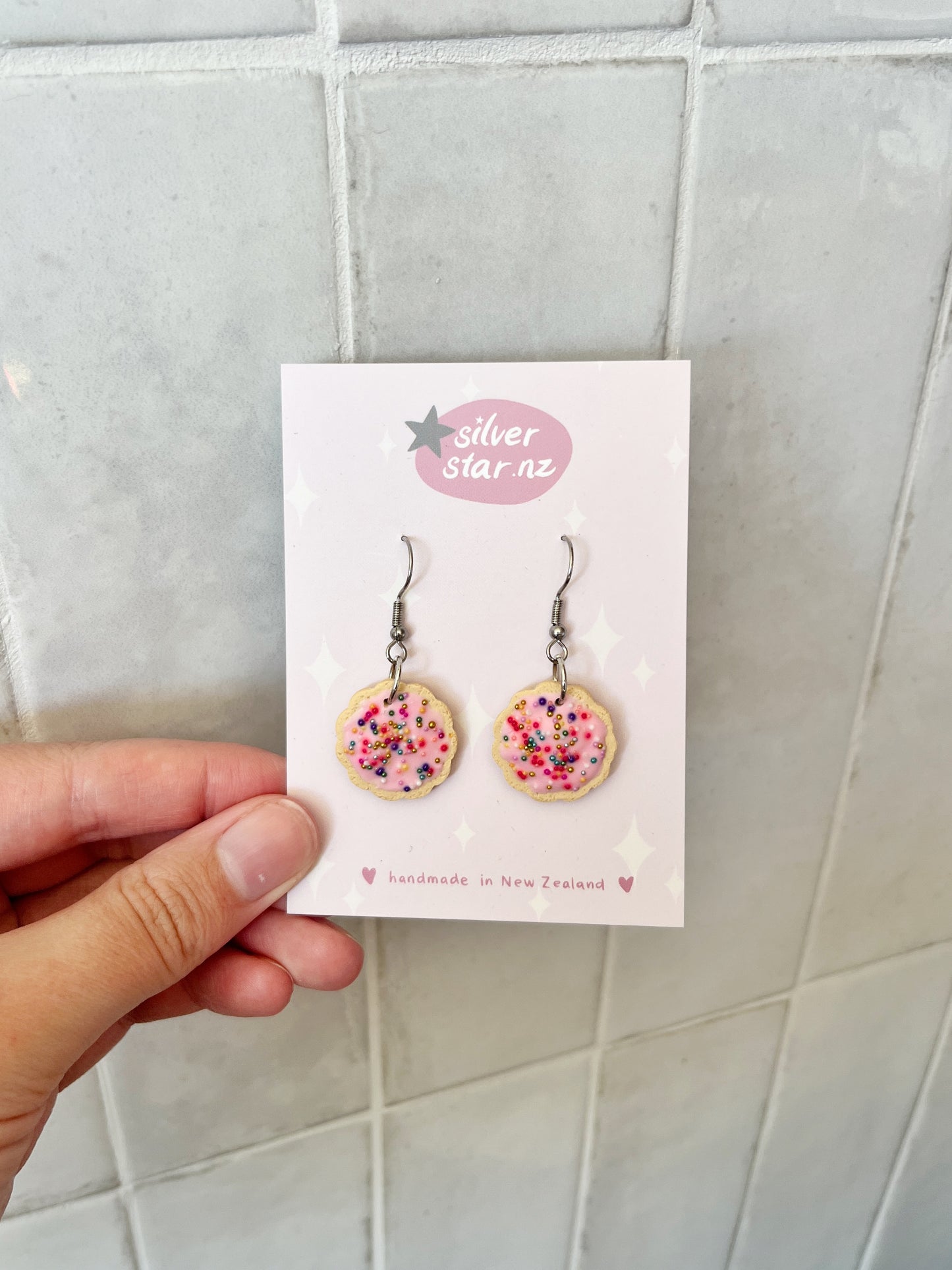 A hand holds a card displaying silverstar.nz Hundreds and Thousands Biscuit Earrings—handmade polymer clay earrings shaped like pink frosted cookies with colorful sprinkles, featuring silverstar.nz and handmade in New Zealand on the card.