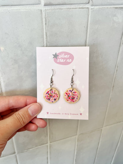 A hand holds a card displaying silverstar.nz Hundreds and Thousands Biscuit Earrings—handmade polymer clay earrings shaped like pink frosted cookies with colorful sprinkles, featuring silverstar.nz and handmade in New Zealand on the card.