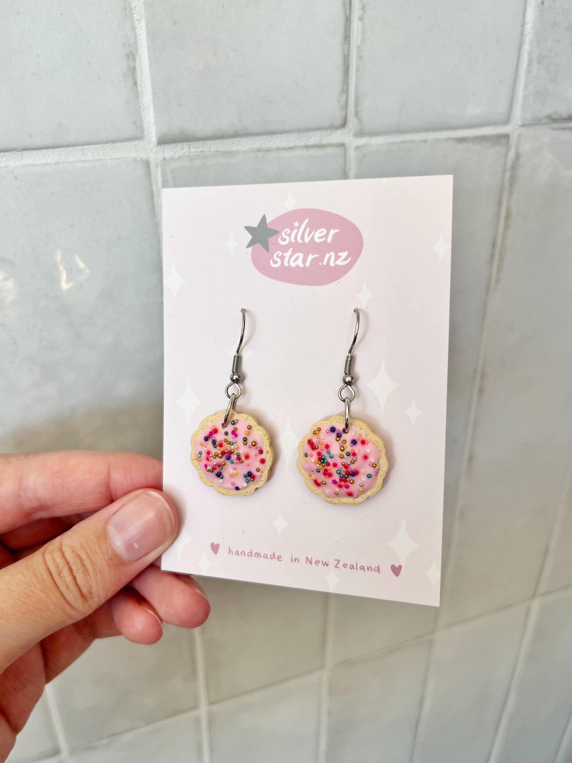 A hand holds a card displaying Hundreds and Thousands Biscuit Earrings by silverstar.nz—handmade polymer clay earrings shaped like pink-frosted cookies with colorful sprinkles, featuring stainless steel hooks against a tiled backdrop.