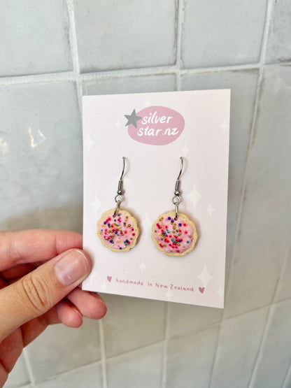 A hand holds a card displaying Hundreds and Thousands Biscuit Earrings by silverstar.nz—handmade polymer clay earrings shaped like pink-frosted cookies with colorful sprinkles, featuring stainless steel hooks against a tiled backdrop.