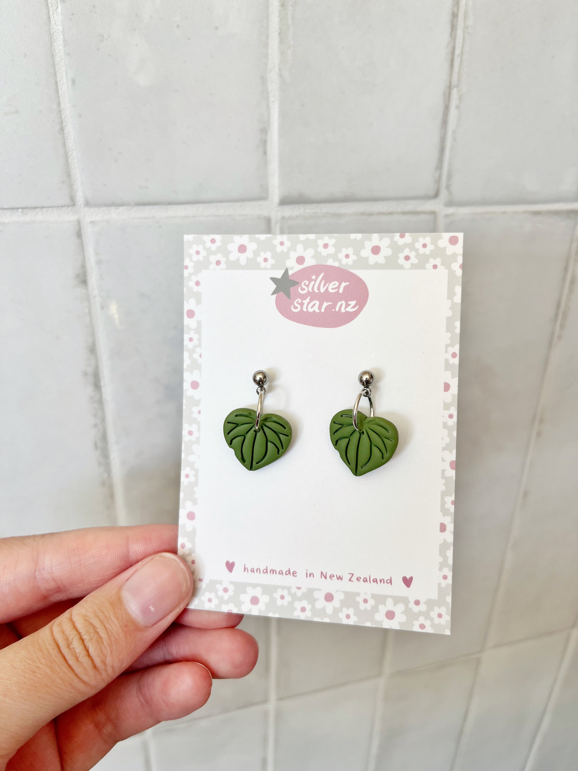 A hand holds a card showing Kawakawa Polymer Clay Earrings with silver studs. The card features the silverstar.nz brand, reads handmade in New Zealand, and has a floral border against a tiled background.