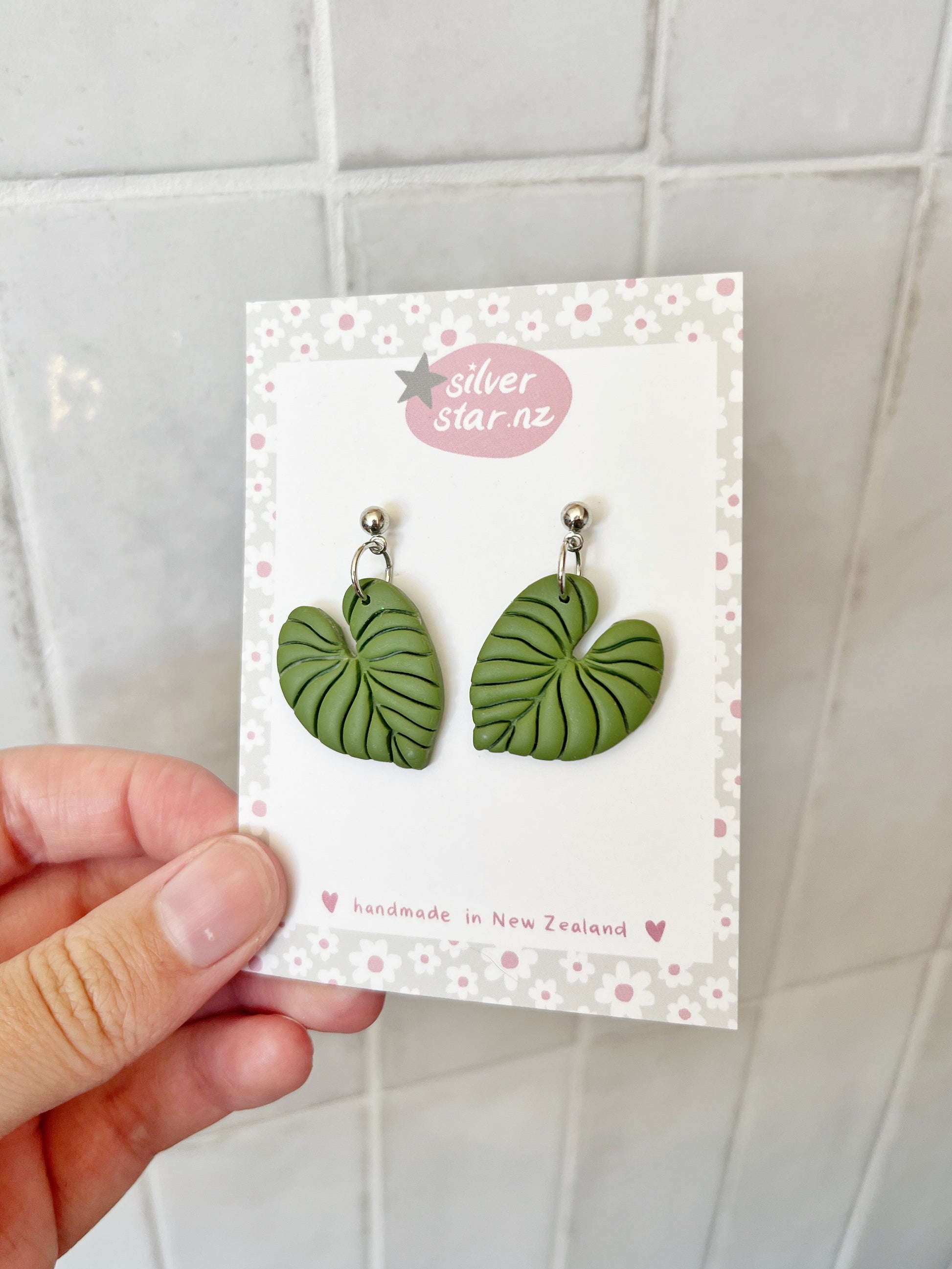 A hand holds a card showing Heart Leaf Polymer Clay Earrings with veined green details by silverstar.nz, handmade in New Zealand. The card features a floral border, and a white tiled wall in the background highlights their unique style.