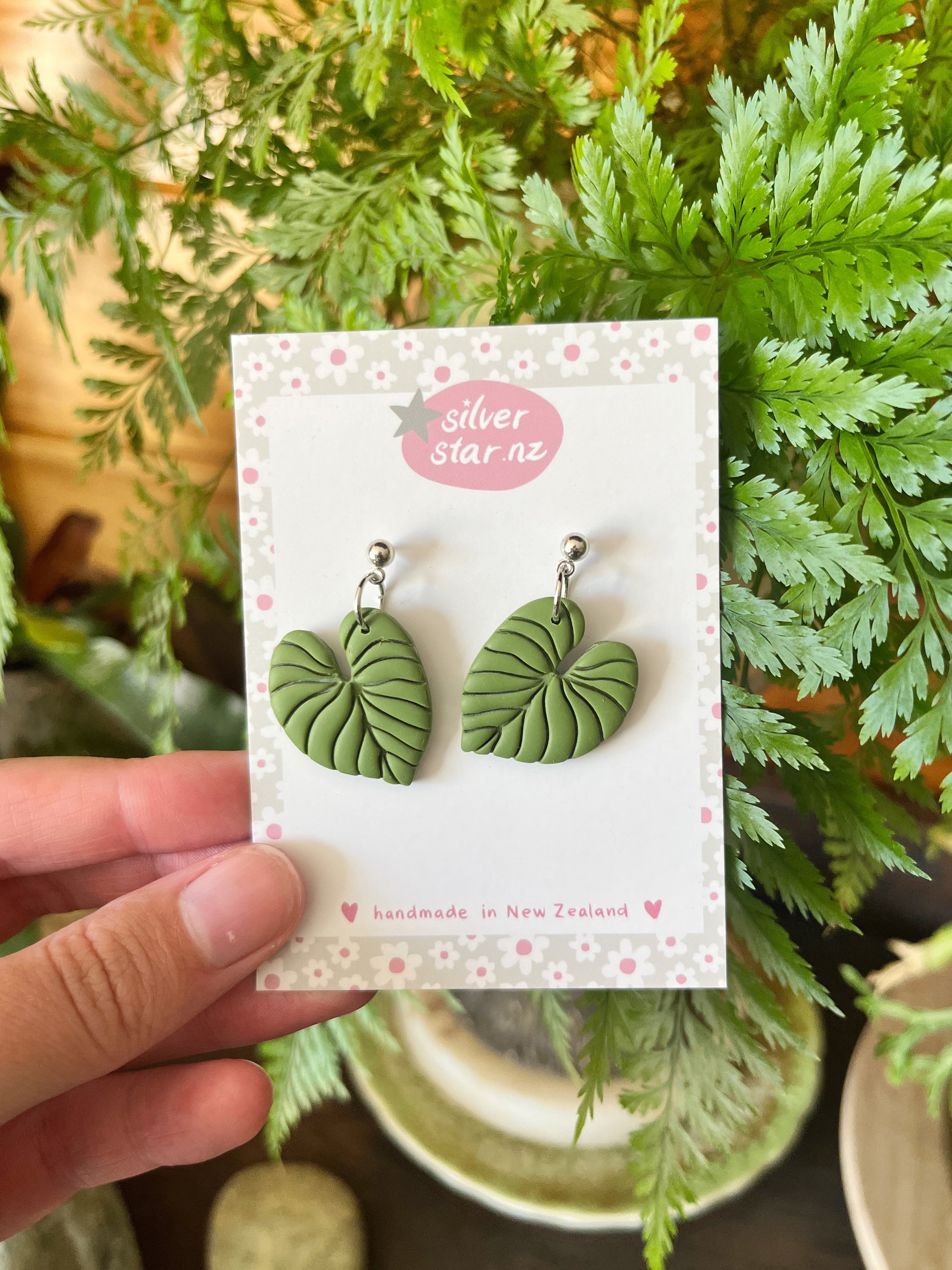 A hand holds a card labeled silverstar.nz, showing Heart Leaf Polymer Clay Earrings with green leaf shapes and silver studs. The card says handmade in New Zealand, with green leafy plants blurred in the background.