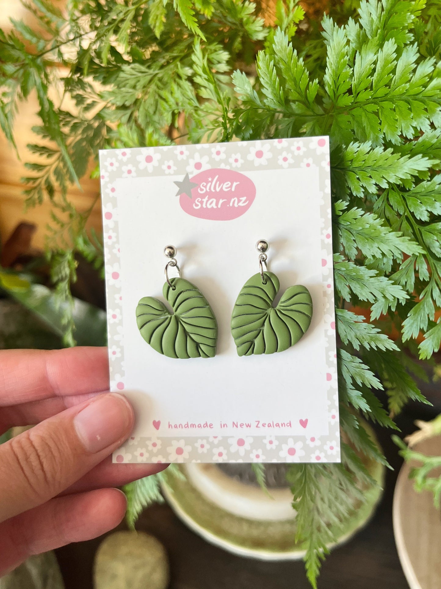 A hand holds a card displaying Heart Leaf Polymer Clay Earrings by silverstar.nz, labeled “handmade in New Zealand.” Green foliage in the background highlights the earrings unique design.