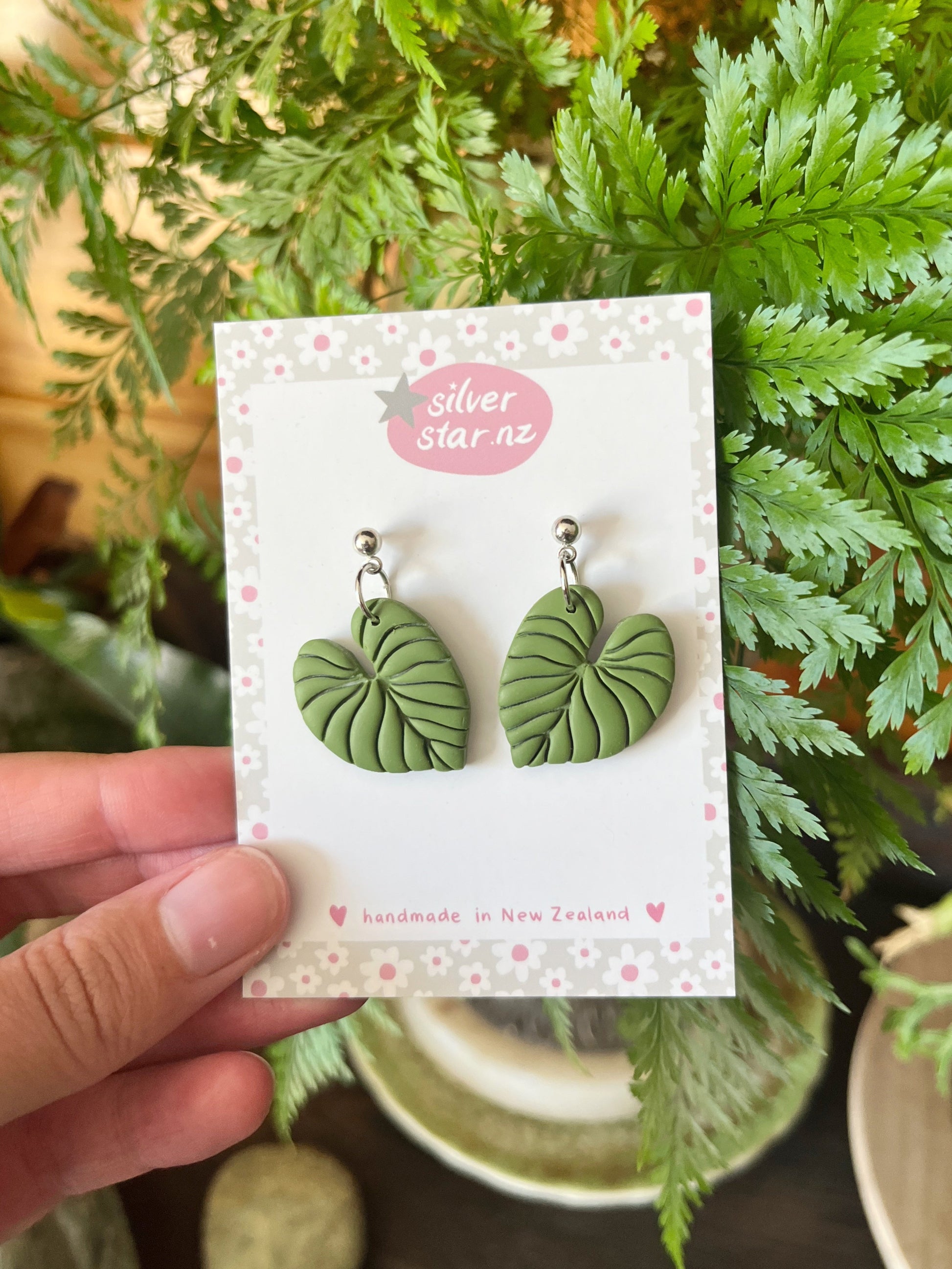 A hand holds a card displaying Heart Leaf Polymer Clay Earrings by silverstar.nz, labeled “handmade in New Zealand.” Green foliage in the background highlights the earrings unique design.