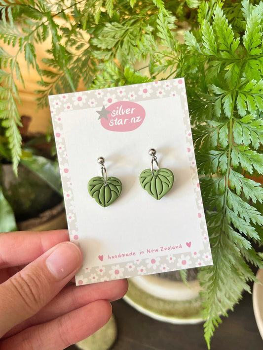 A hand holds a card displaying Kawakawa Polymer Clay Earrings with stainless steel hooks. The card reads silverstar.nz and handmade in New Zealand, set against a background of lush green plants.