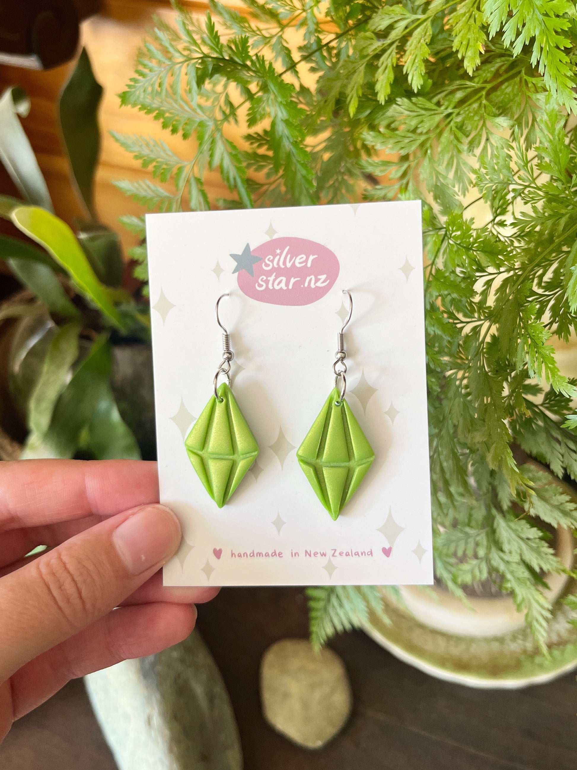 A hand holds a card labeled silverstar.nz, handmade in New Zealand, displaying Sims Plumbob Polymer Clay Earrings—green, diamond-shaped with silver hooks—set against green plants and stones.