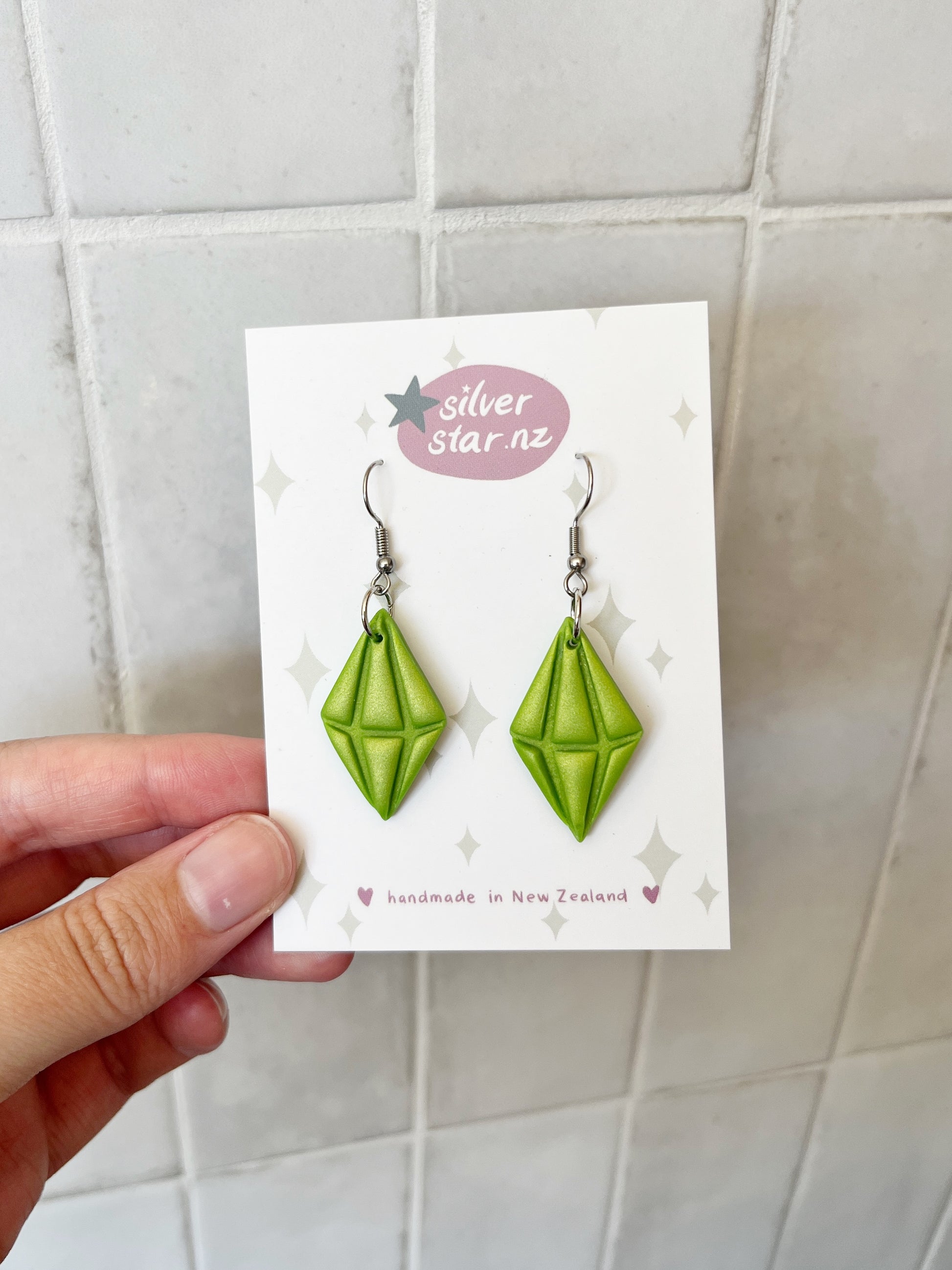 A hand displays a card with Sims Plumbob Polymer Clay Earrings—green, diamond-shaped, inspired by The Sims. The card says silverstar.nz and handmade in New Zealand, set against a tiled background.