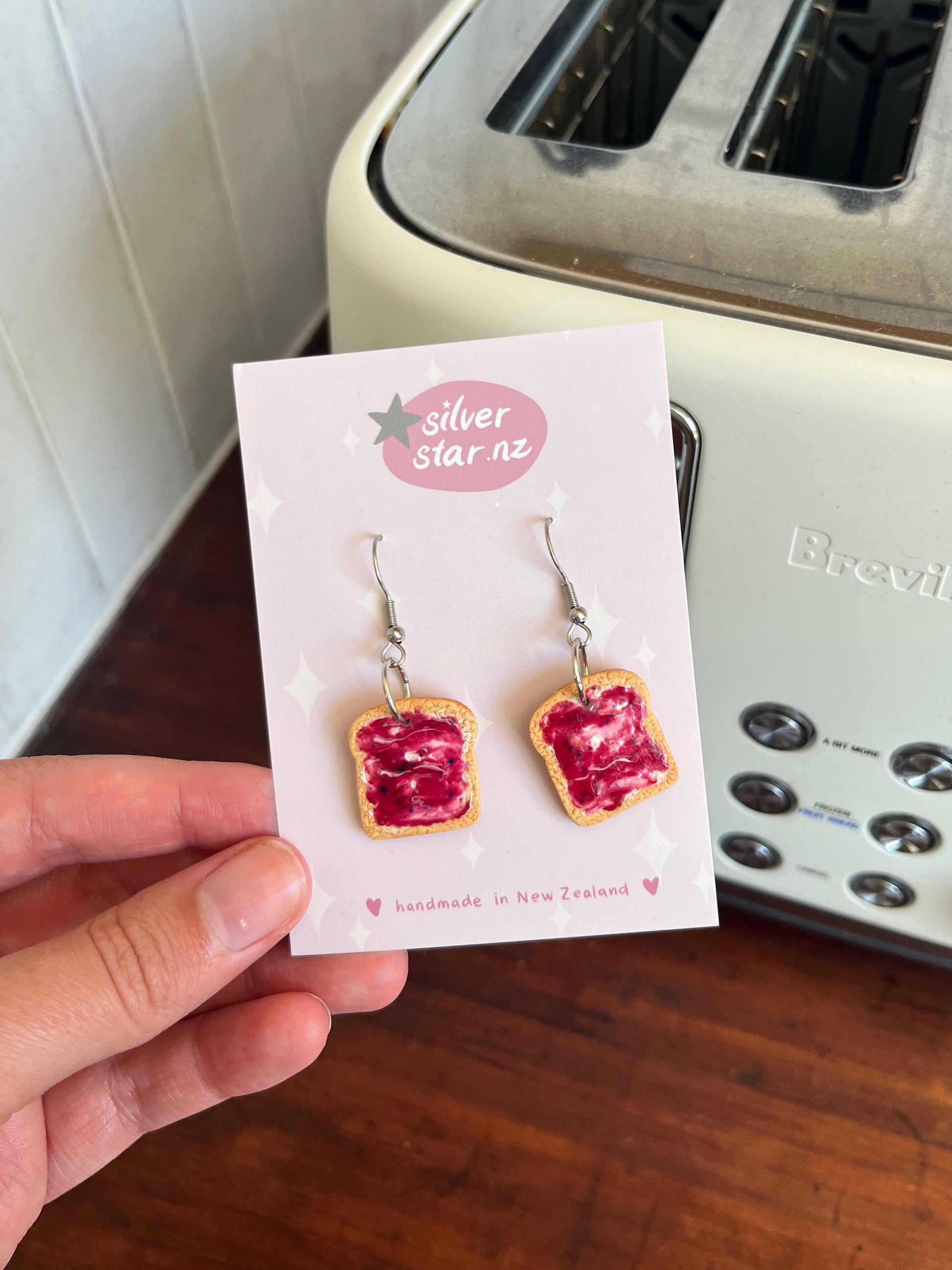 A hand holds Berry Jam on Toast Polymer Clay Earrings by silverstar.nz, displayed on a branded card before a white Breville toaster. These handmade earrings feature toast with red jam and dangle from stainless steel hooks.
