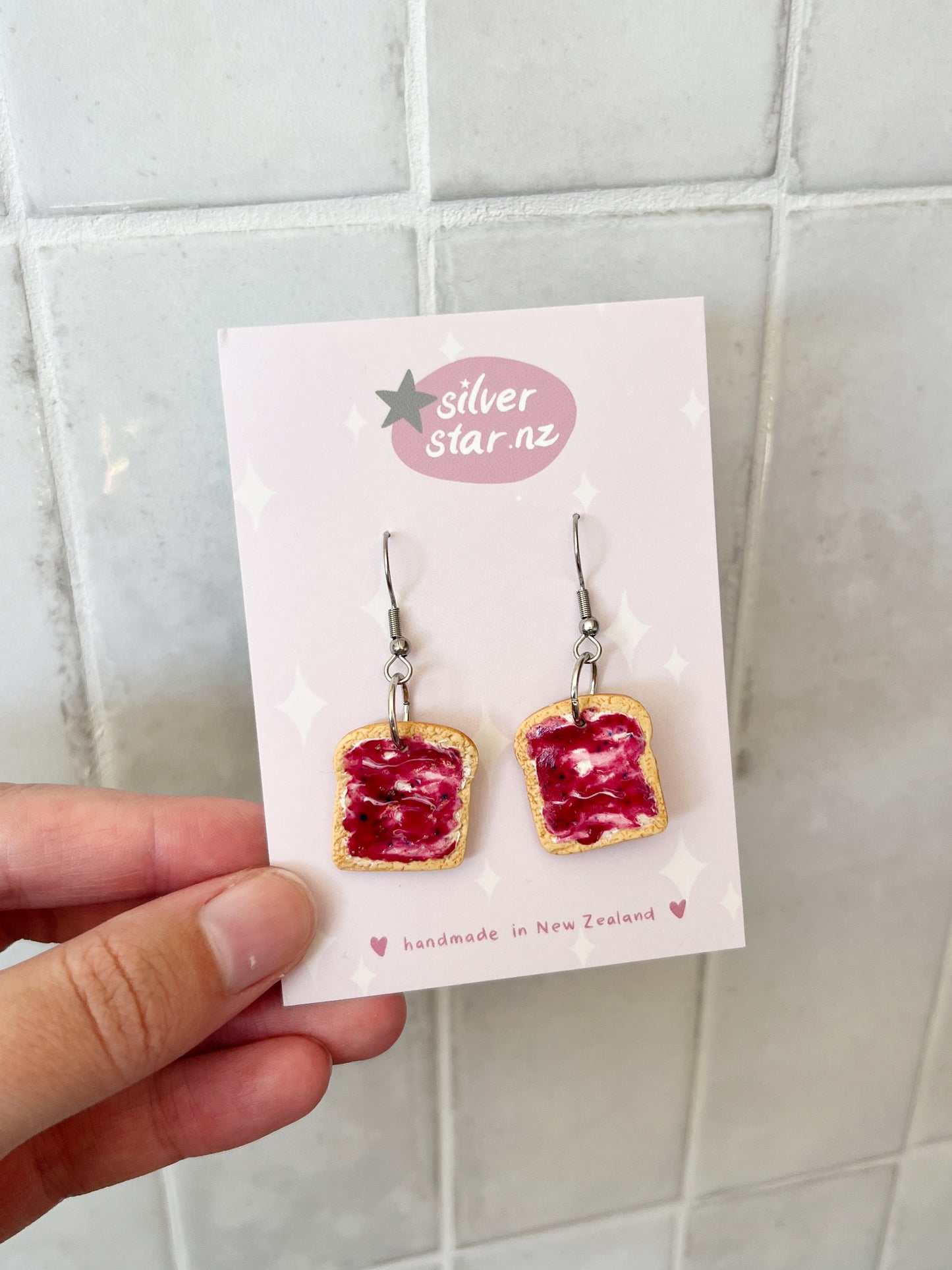 A hand holds a card from silverstar.nz displaying Berry Jam on Toast Polymer Clay Earrings with stainless steel hooks, set against a tiled background. The card says handmade in New Zealand.
