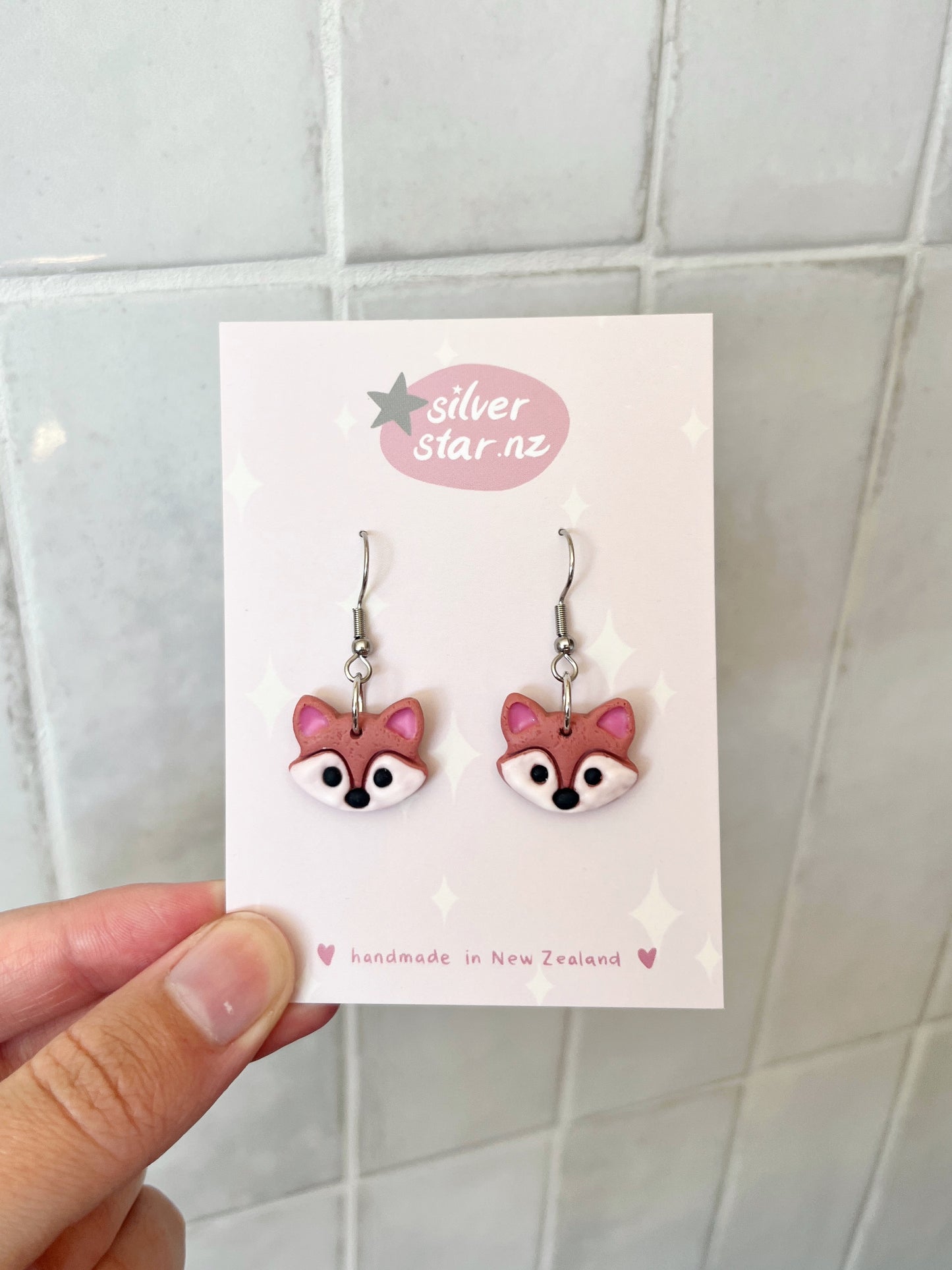 A hand holds a card featuring two handmade polymer clay earrings shaped like pink fox faces, with silverstar.nz at the top and handmade in New Zealand at the bottom. The funky designs hang on stainless steel hooks against a tiled background. Brand: silverstar.nz; Product: Fox Polymer Clay Earrings.