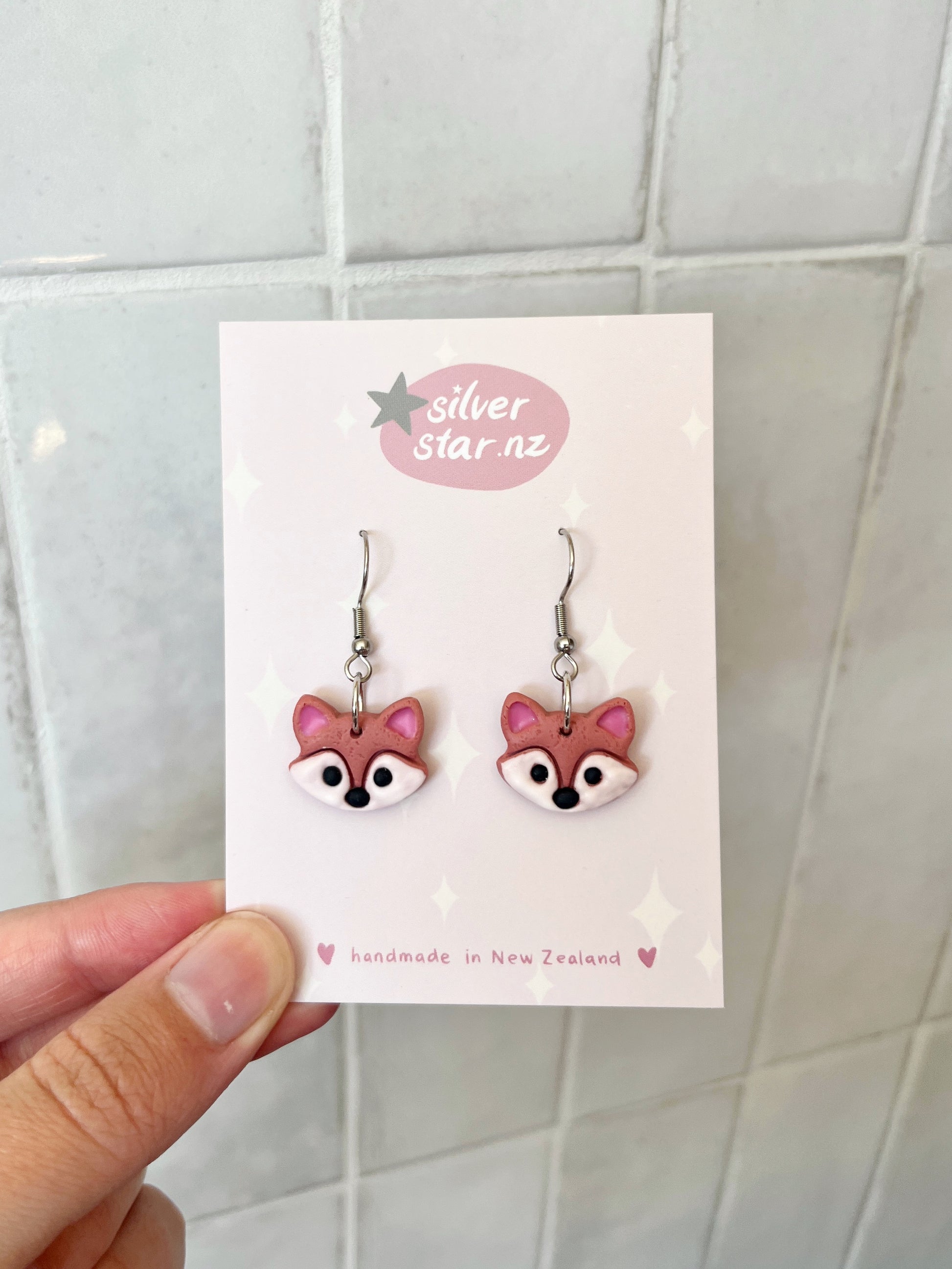 A hand holds a card featuring two handmade polymer clay earrings shaped like pink fox faces, with silverstar.nz at the top and handmade in New Zealand at the bottom. The funky designs hang on stainless steel hooks against a tiled background. Brand: silverstar.nz; Product: Fox Polymer Clay Earrings.