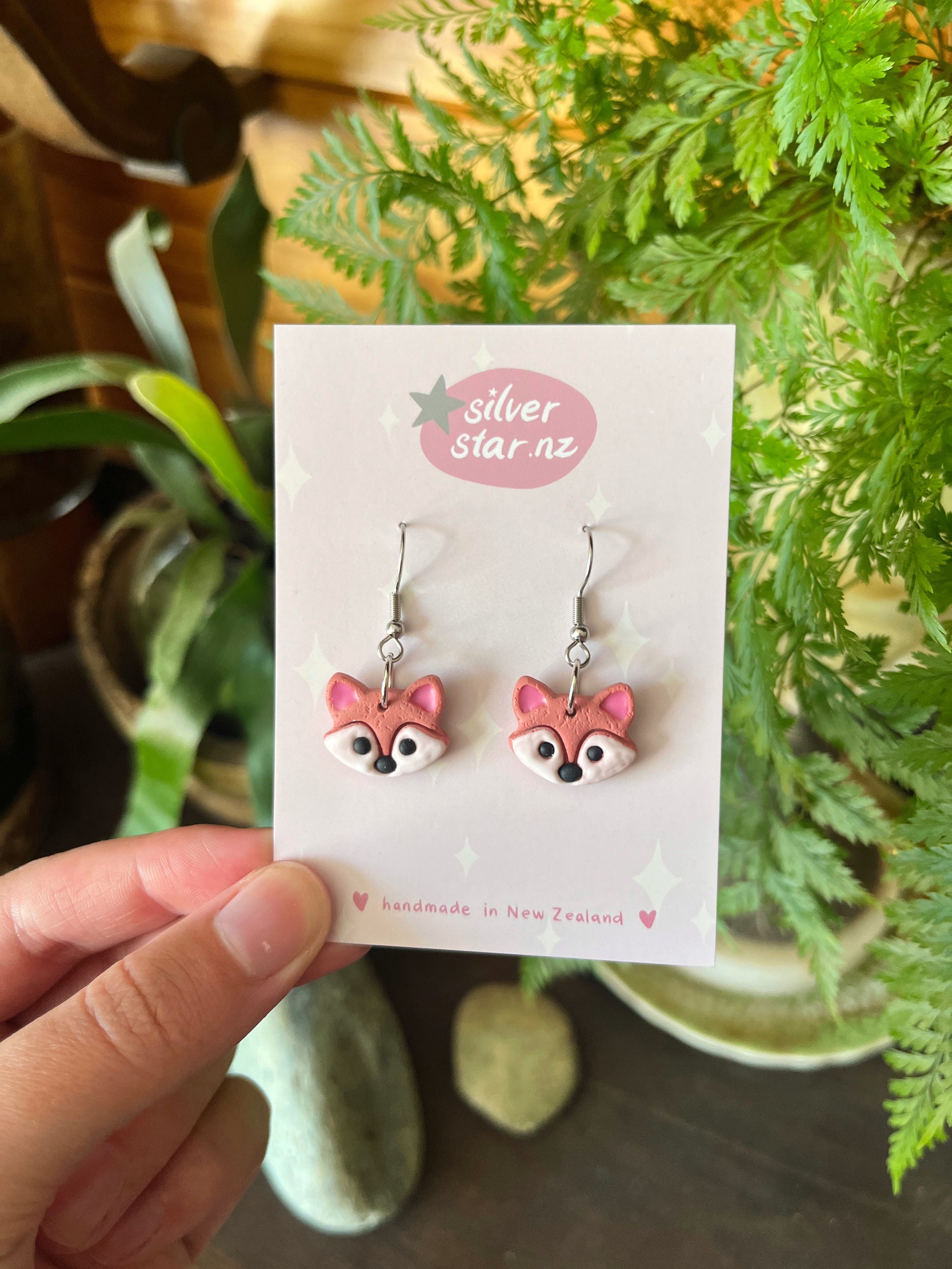 A hand displays pink fox-shaped earrings made from handmade polymer clay on a Silver Star NZ card. Surrounded by green houseplants, stones, and wood, they are labeled as handmade in New Zealand with a unique funky design. Product: Fox Polymer Clay Earrings by silverstar.nz.