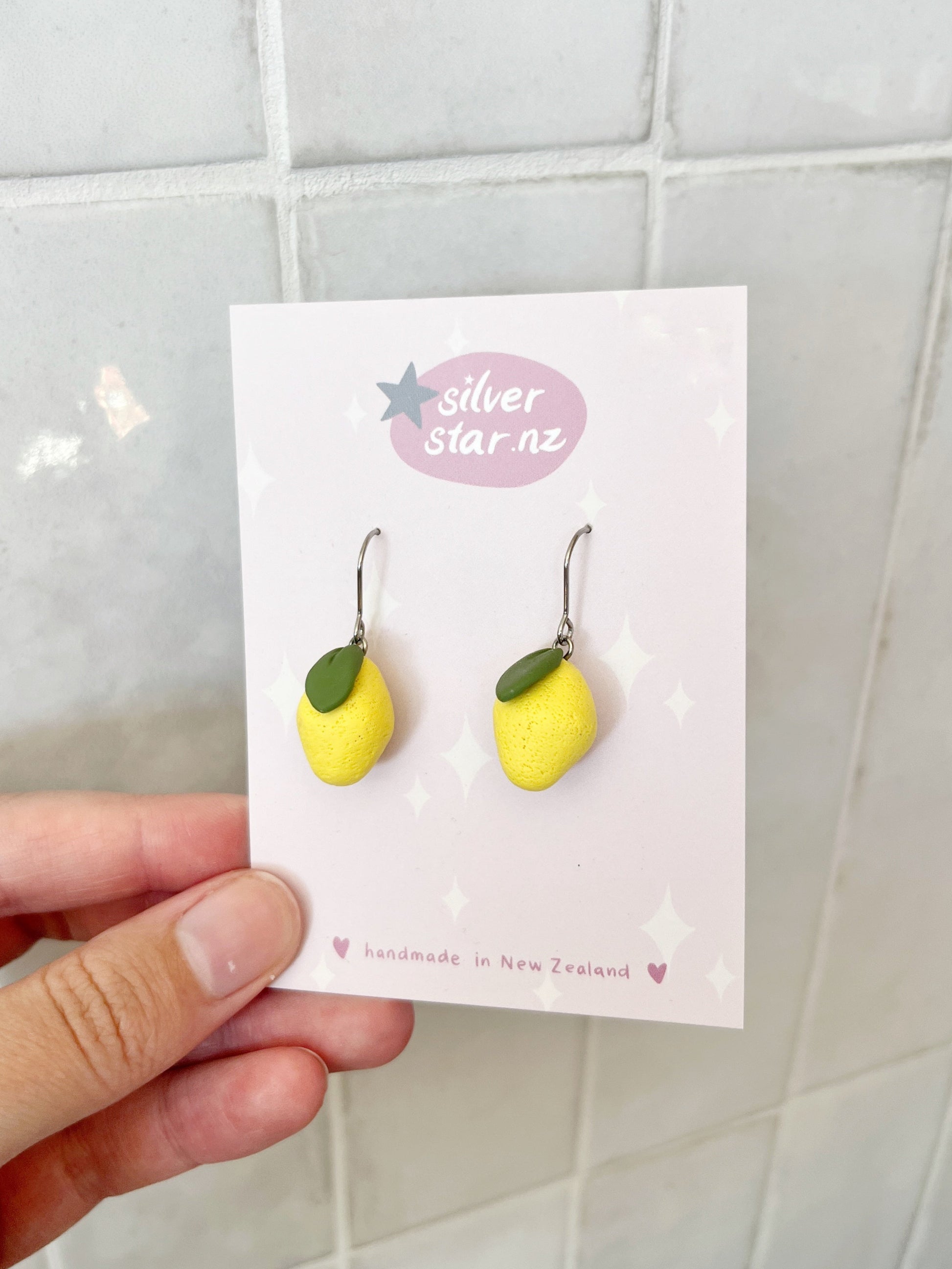 A hand displays a card featuring Mini Lemon Polymer Clay Earrings, shaped like yellow lemons with tiny green leaves, on a tiled background. The card reads silverstar.nz and handmade in New Zealand.