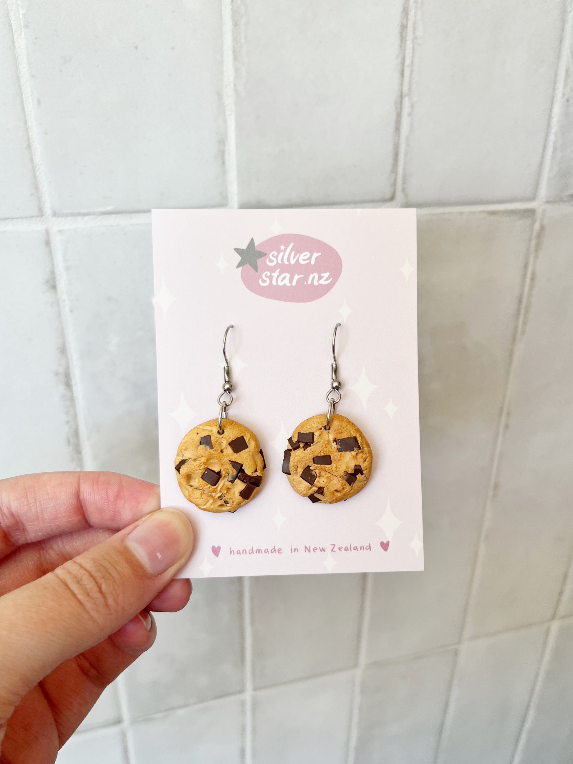 A hand displays a card with artisan-crafted Chocolate Chip Cookie Earrings by silverstar.nz, made from polymer clay with black star motifs on a light background. The card reads handmade in New Zealand, all set against a tiled wall for contrast.