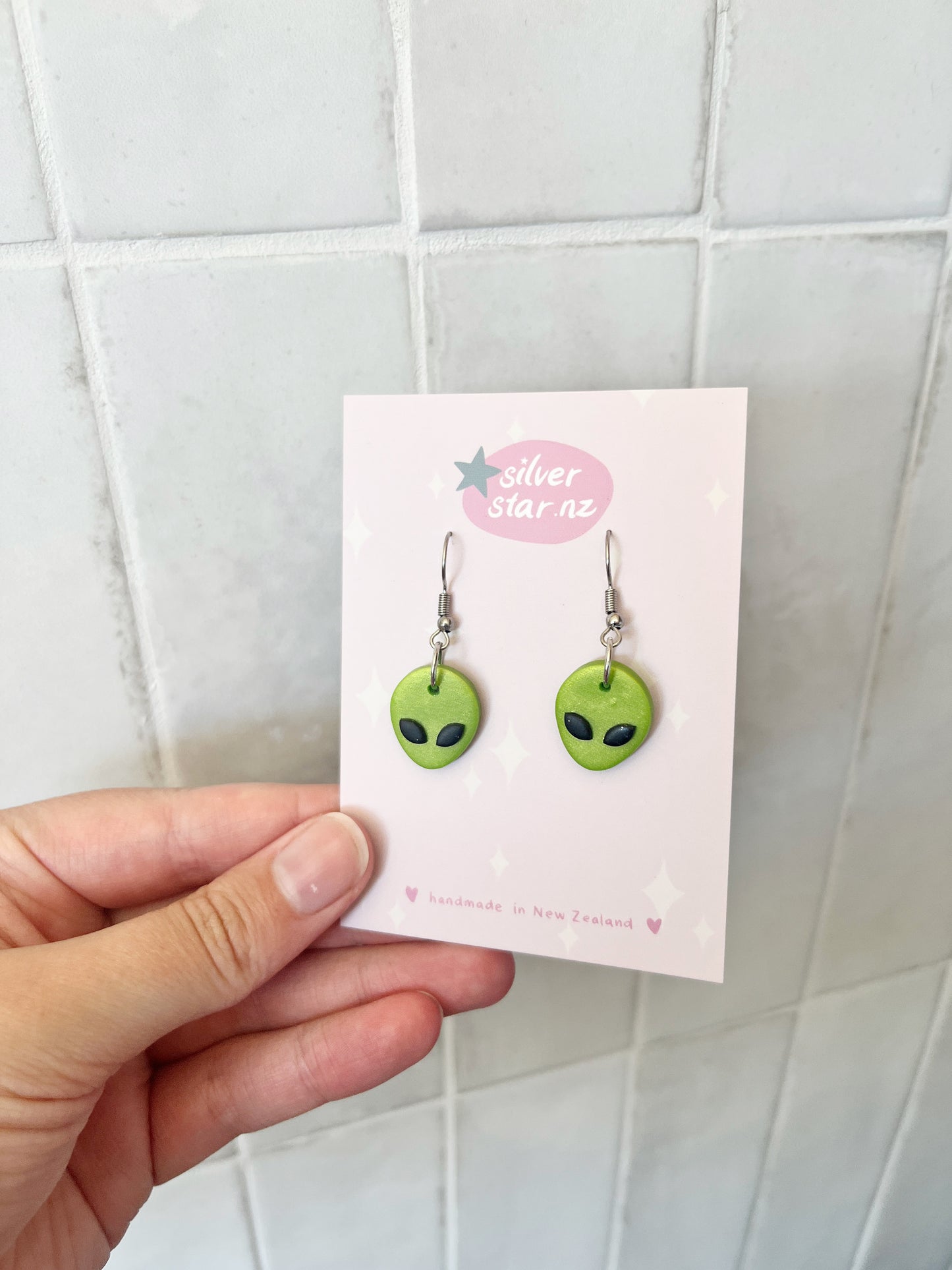 A hand holds a card featuring Alien Polymer Clay Earrings shaped like green alien faces with black eyes. The card shows the brand silverstar.nz and states they are handmade in New Zealand, set against a tiled background.