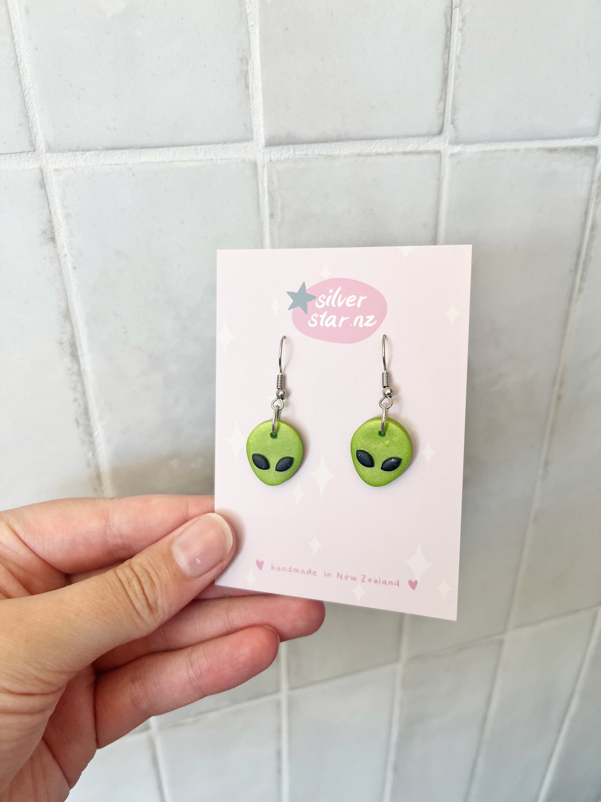 A hand holds a card featuring Alien Polymer Clay Earrings shaped like green alien faces with black eyes. The card shows the brand silverstar.nz and states they are handmade in New Zealand, set against a tiled background.