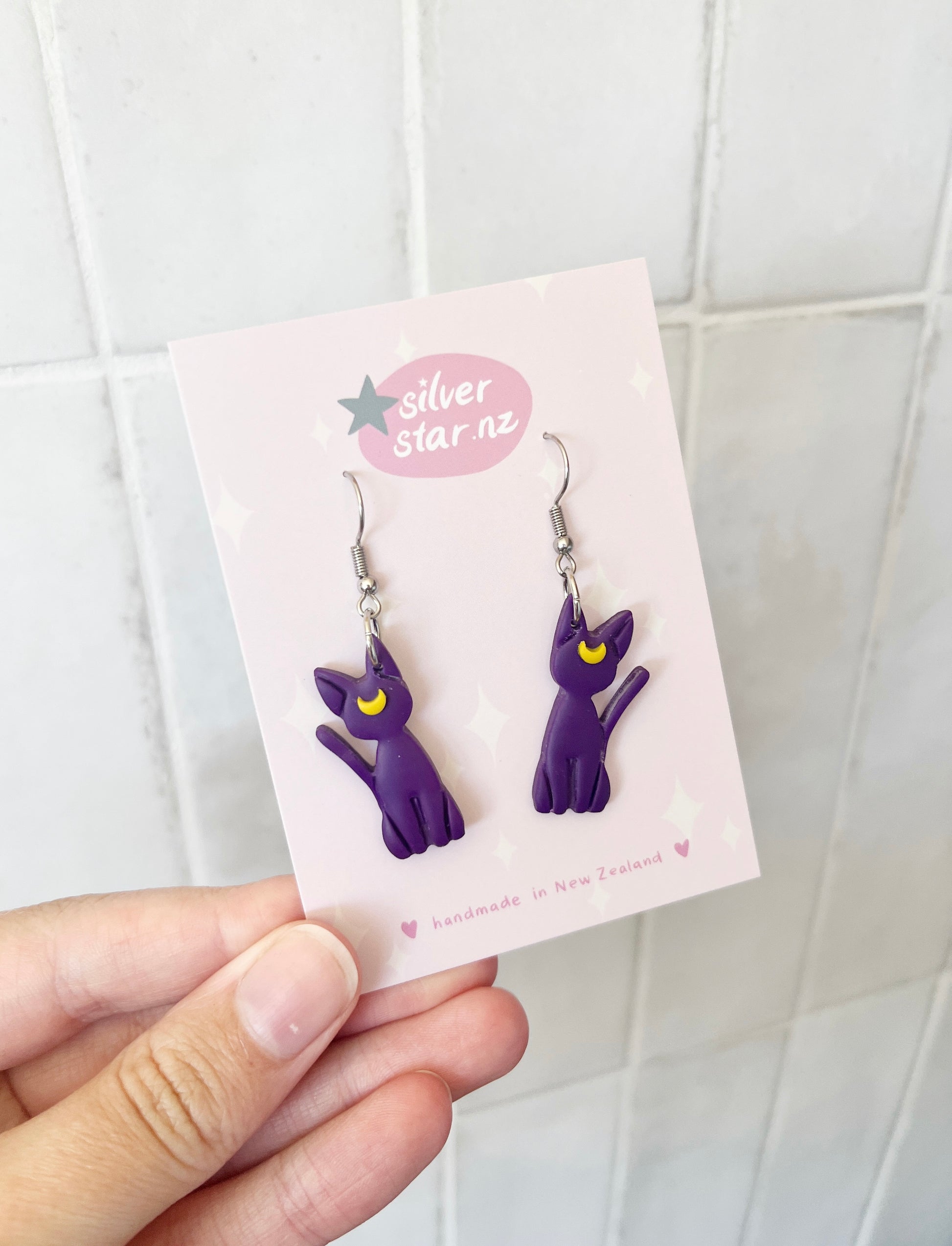 A hand showcases Luna Cat Polymer Clay Earrings: purple cat and crescent moon design on stainless steel hooks, presented on a pink silverstar.nz card with stars against a tiled background.