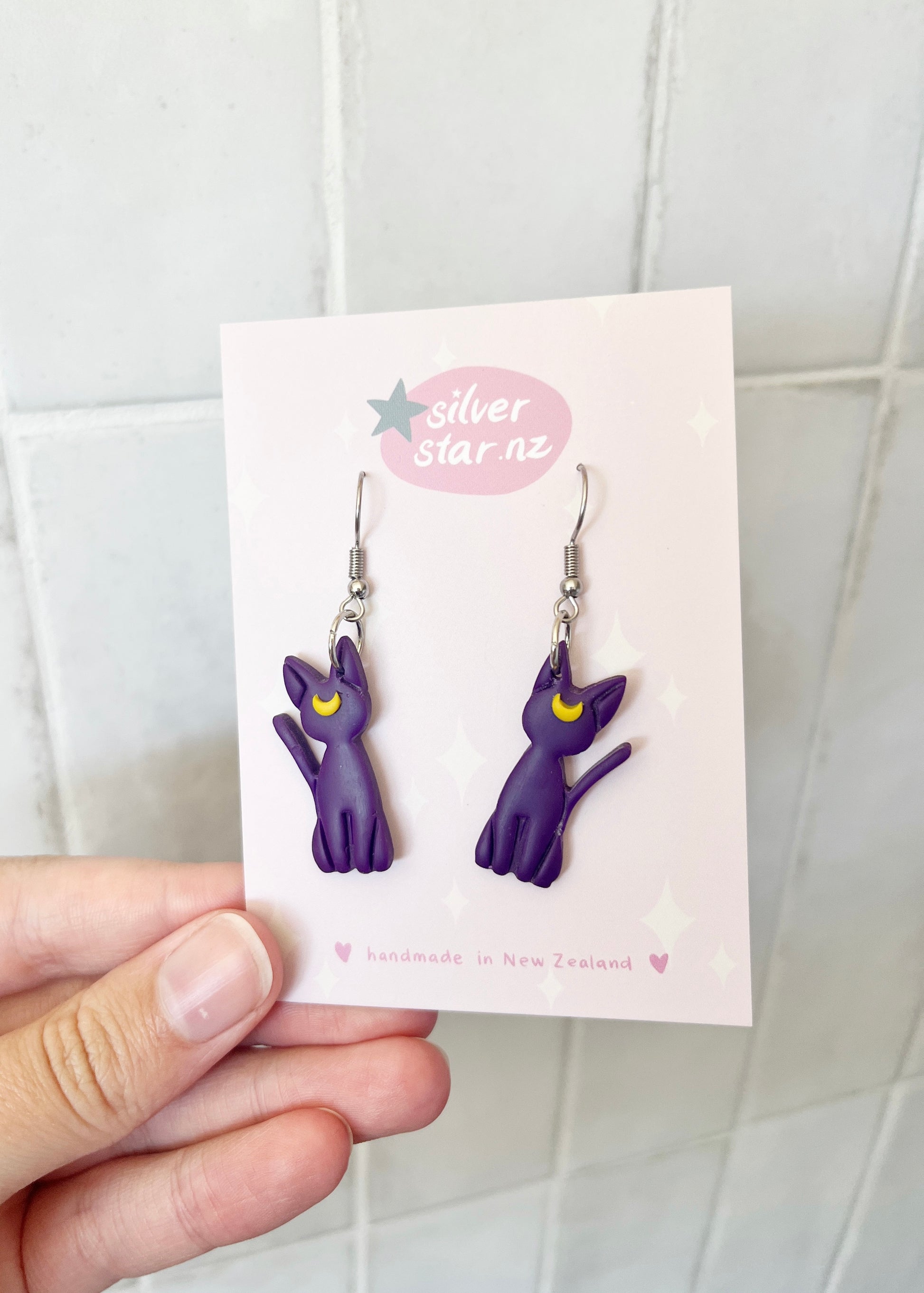 These unique, funky purple Luna Cat earrings by silverstar.nz feature cat shapes with yellow moon details, crafted from handmade polymer clay and set on stainless steel hooks. Displayed on a card that reads handmade in New Zealand against a light tiled background.