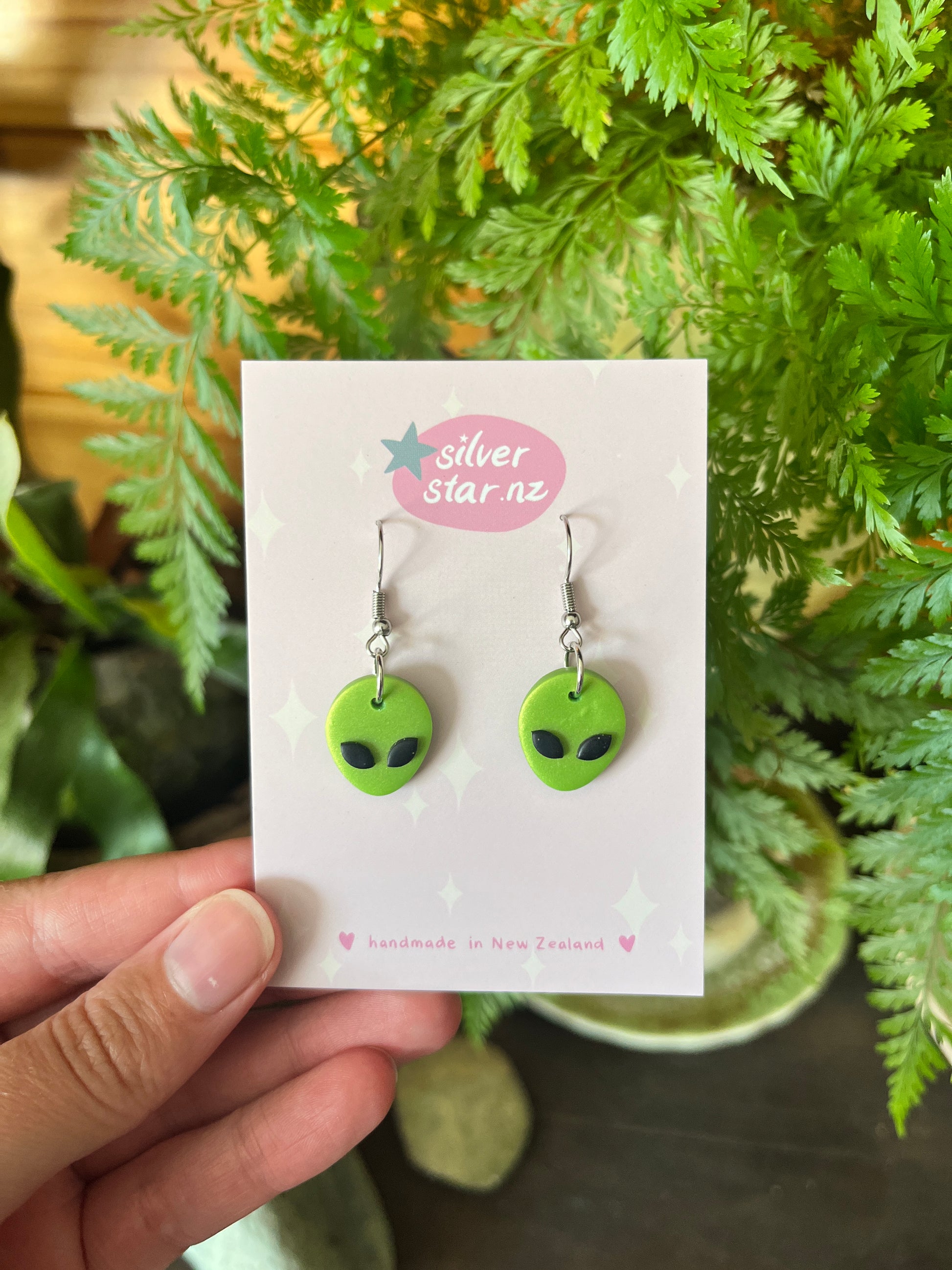 A hand holds a card from silverstar.nz displaying Alien Polymer Clay Earrings shaped like green alien faces with black eyes. The card reads handmade in New Zealand, with ferns and greenery visible in the background.