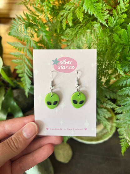 A hand holds a card from silverstar.nz displaying Alien Polymer Clay Earrings shaped like green alien faces with black eyes. The card reads handmade in New Zealand, with ferns and greenery visible in the background.