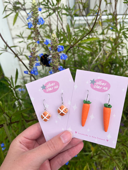 A hand holds cards displaying earrings: one pair is shaped like hot cross buns, the other like carrots. A flowering plant with tiny blue flowers and a bee adds charm. The cards read silverstar.nz and handmade in New Zealand, featuring stainless steel hooks from Hot Cross Bun Polymer Clay Earrings.