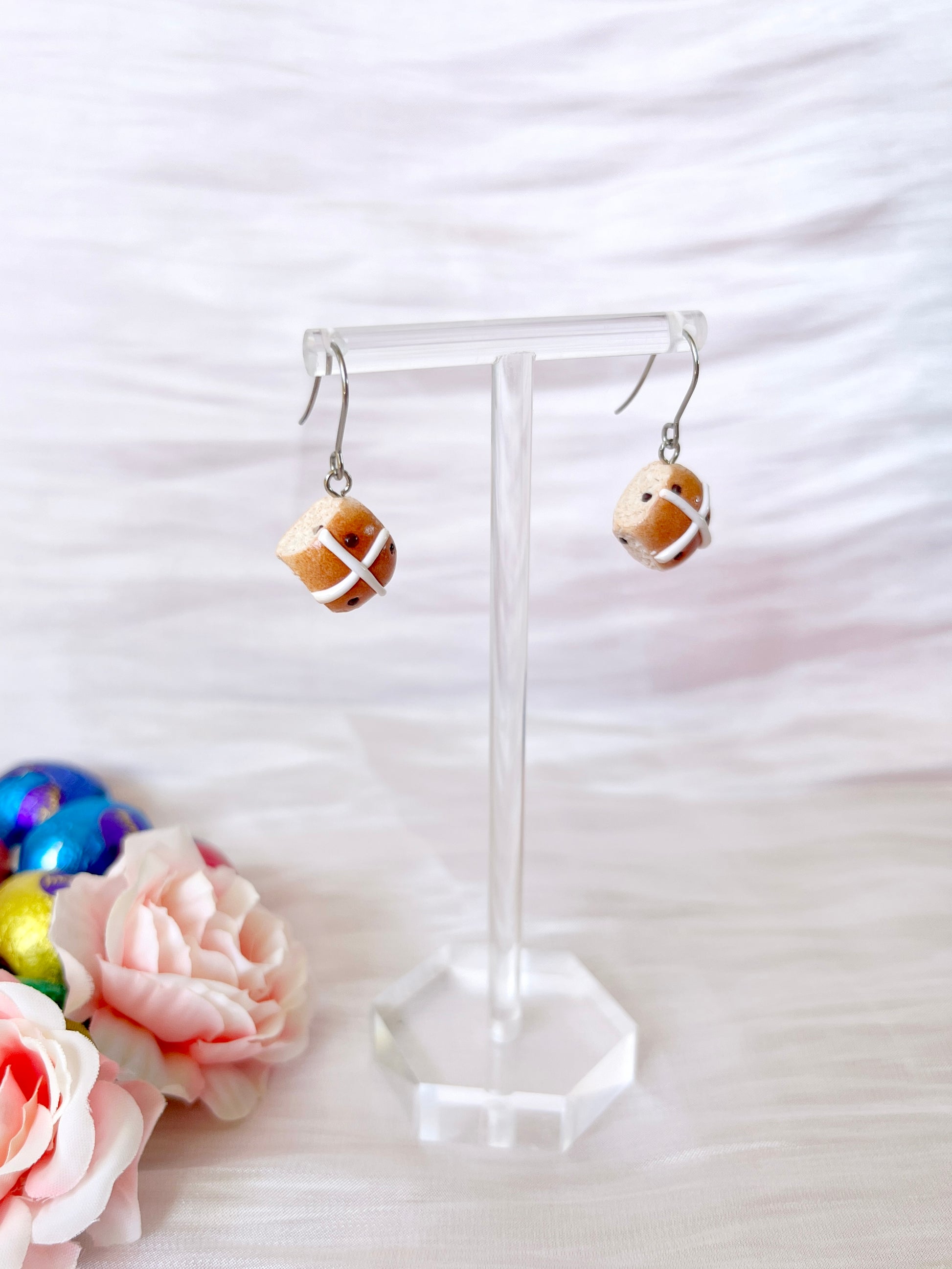 Hot Cross Bun Polymer Clay Earrings by silverstar.nz are handmade in polymer clay with stainless steel hooks, displayed on a clear stand against a soft white fabric. Pink roses and colorful Easter eggs enhance the displays charm.