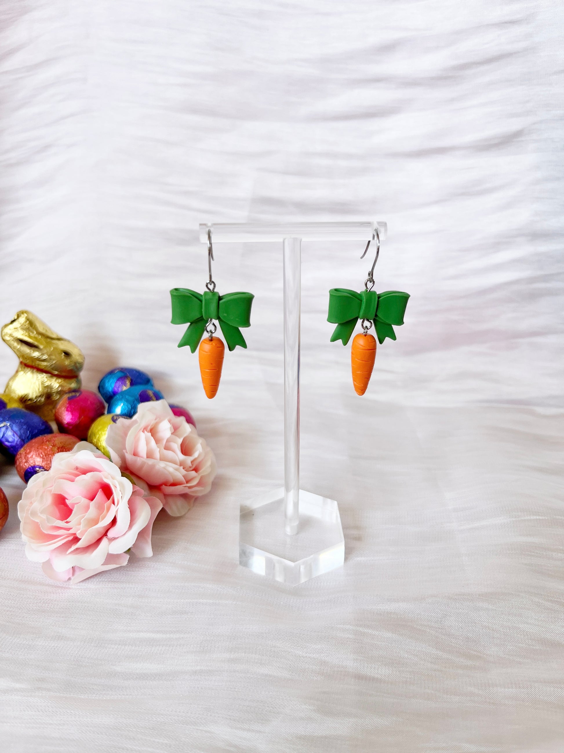 The photo features silverstar.nzs Carrots with a Bow Polymer Clay Earrings on a clear stand, adorned with carrot pendants and green bows. Pink flowers, colorful Easter eggs, and a gold bunny enhance the unique design against a soft white backdrop.