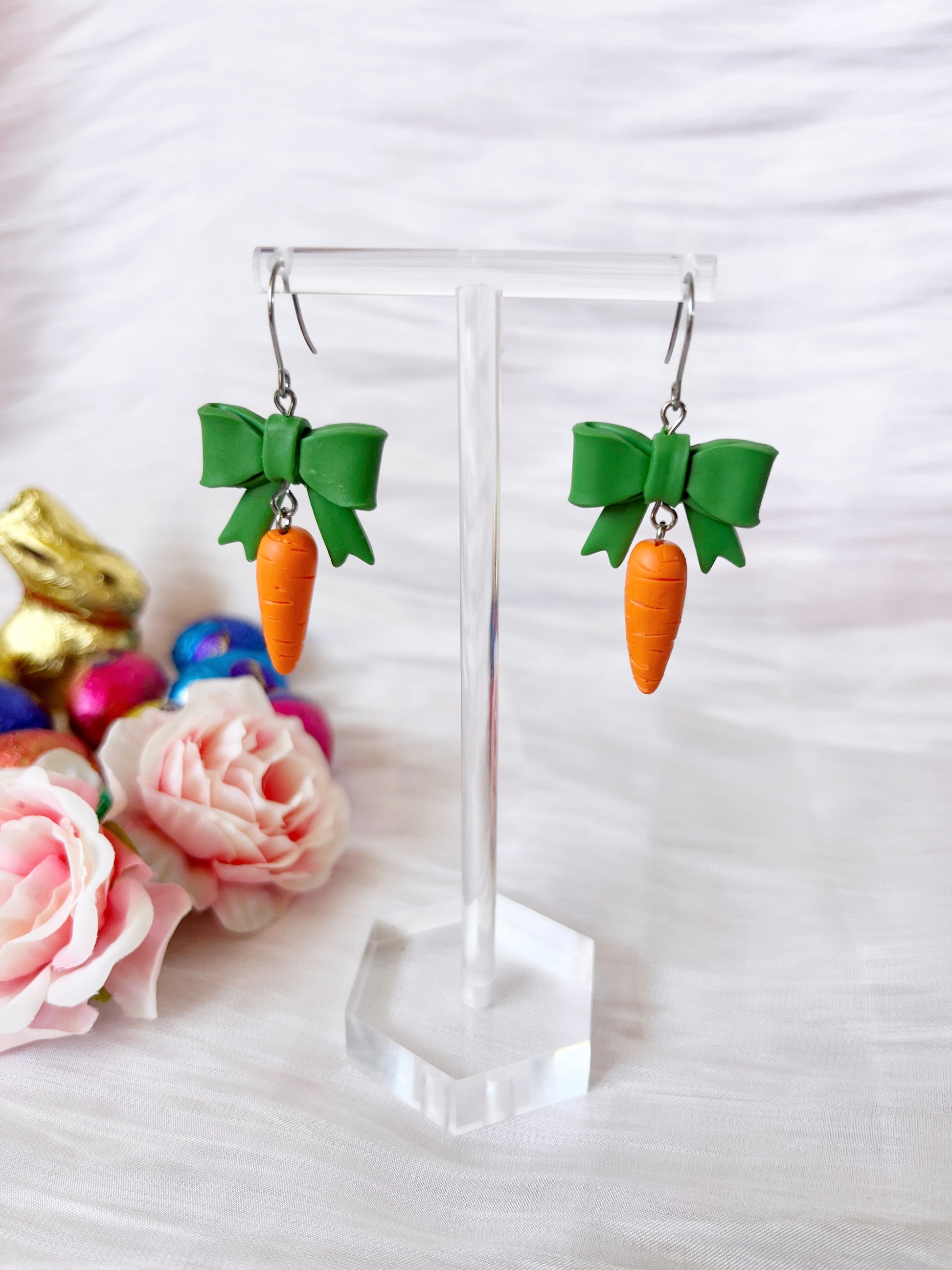 The Carrots with a Bow Polymer Clay Earrings by silverstar.nz, featuring green bows and dangling carrots, are showcased on a clear stand amidst pastel eggs, a gold bunny, and pink roses against soft white fabric.