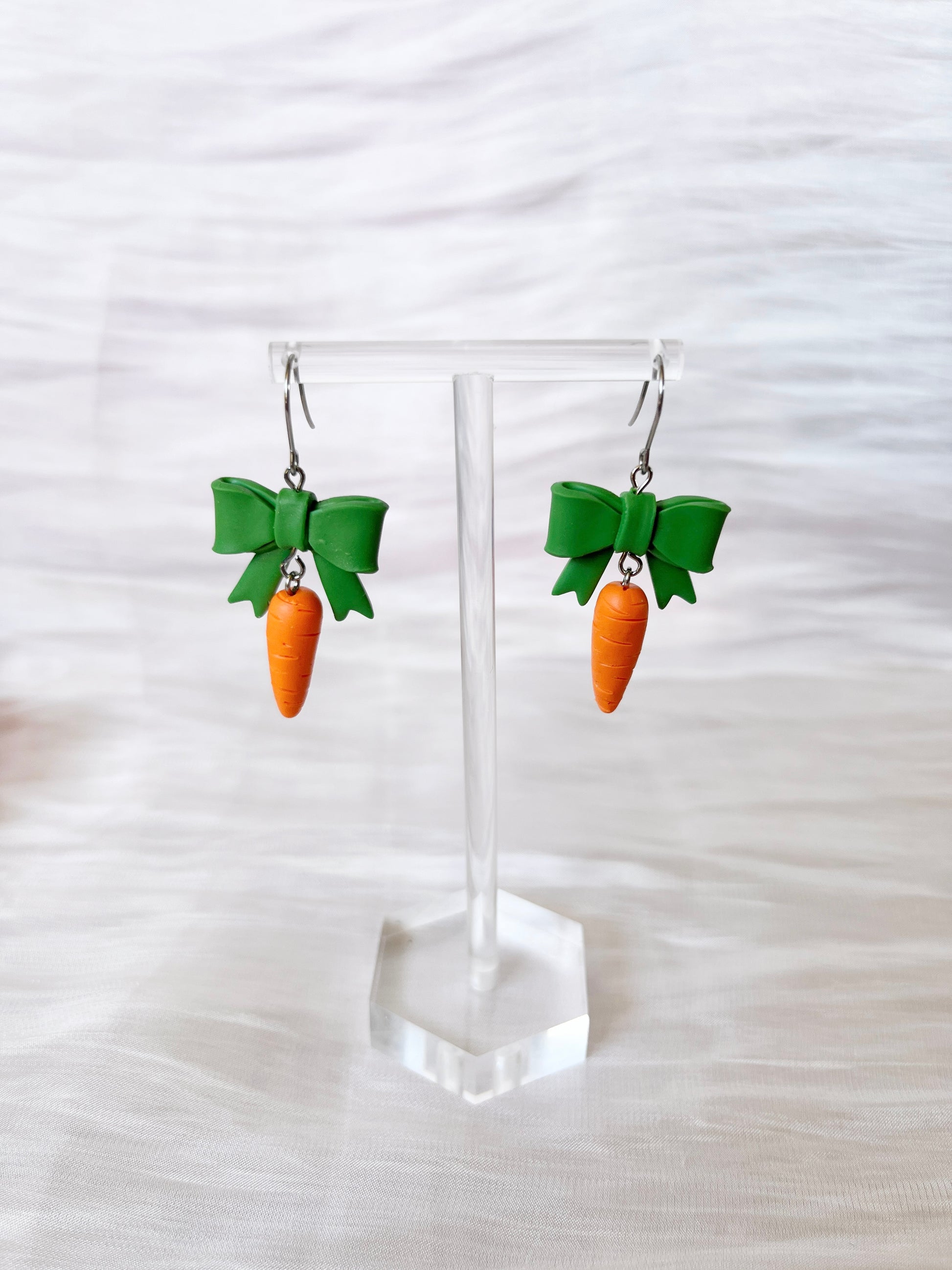 The Carrots with a Bow Polymer Clay Earrings by silverstar.nz feature orange carrot shapes with green tops hanging from silver hoops, complemented by green bow-shaped accents. Displayed on a clear stand against a soft, light background, these earrings offer a unique design.