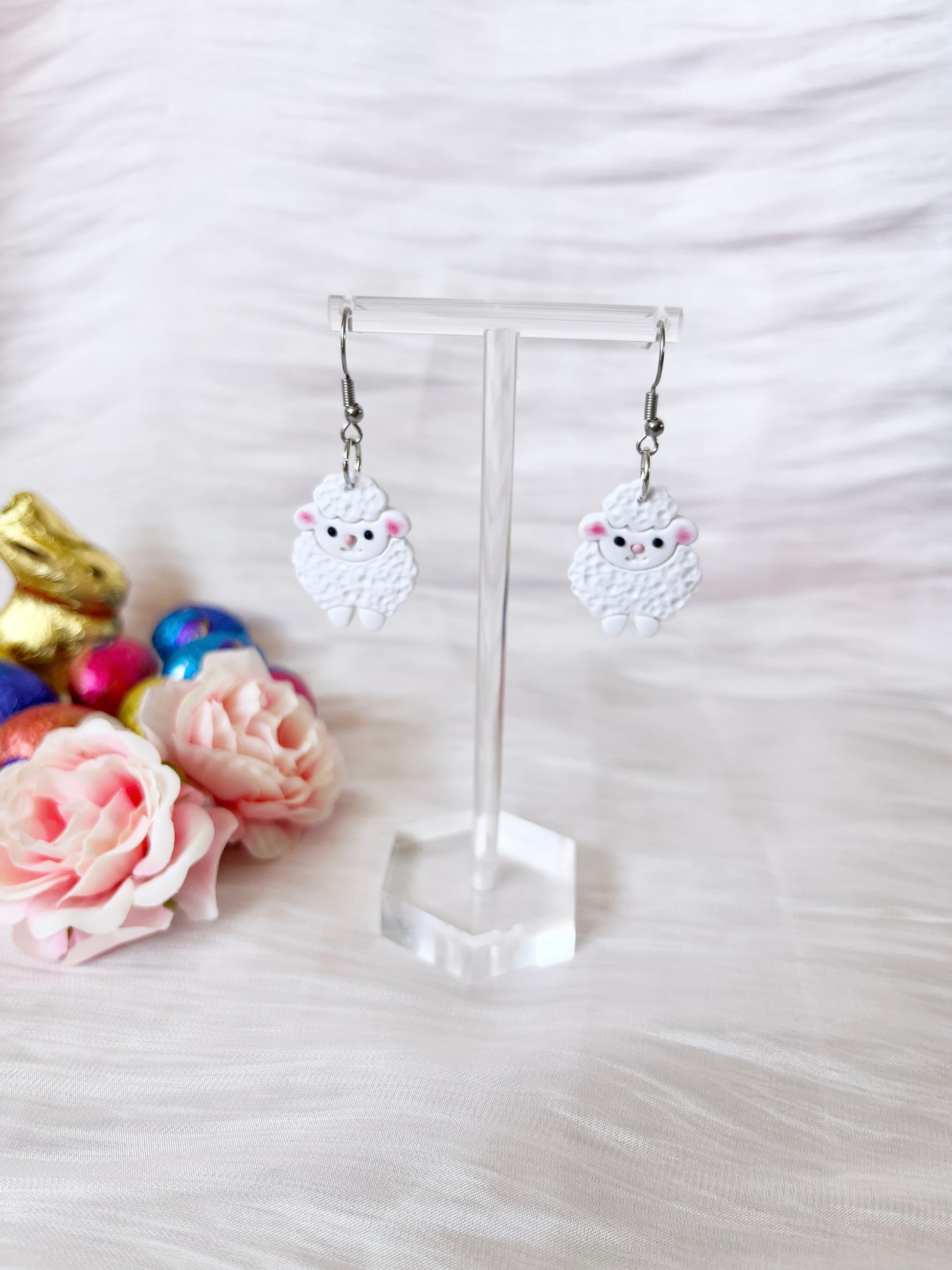 Fluffy Sheep Polymer Clay Earrings by silverstar.nz hang on a clear stand, surrounded by pink roses and colorful foil-wrapped chocolate eggs on soft white fabric. The cute, handmade design adds charm to any outfit.