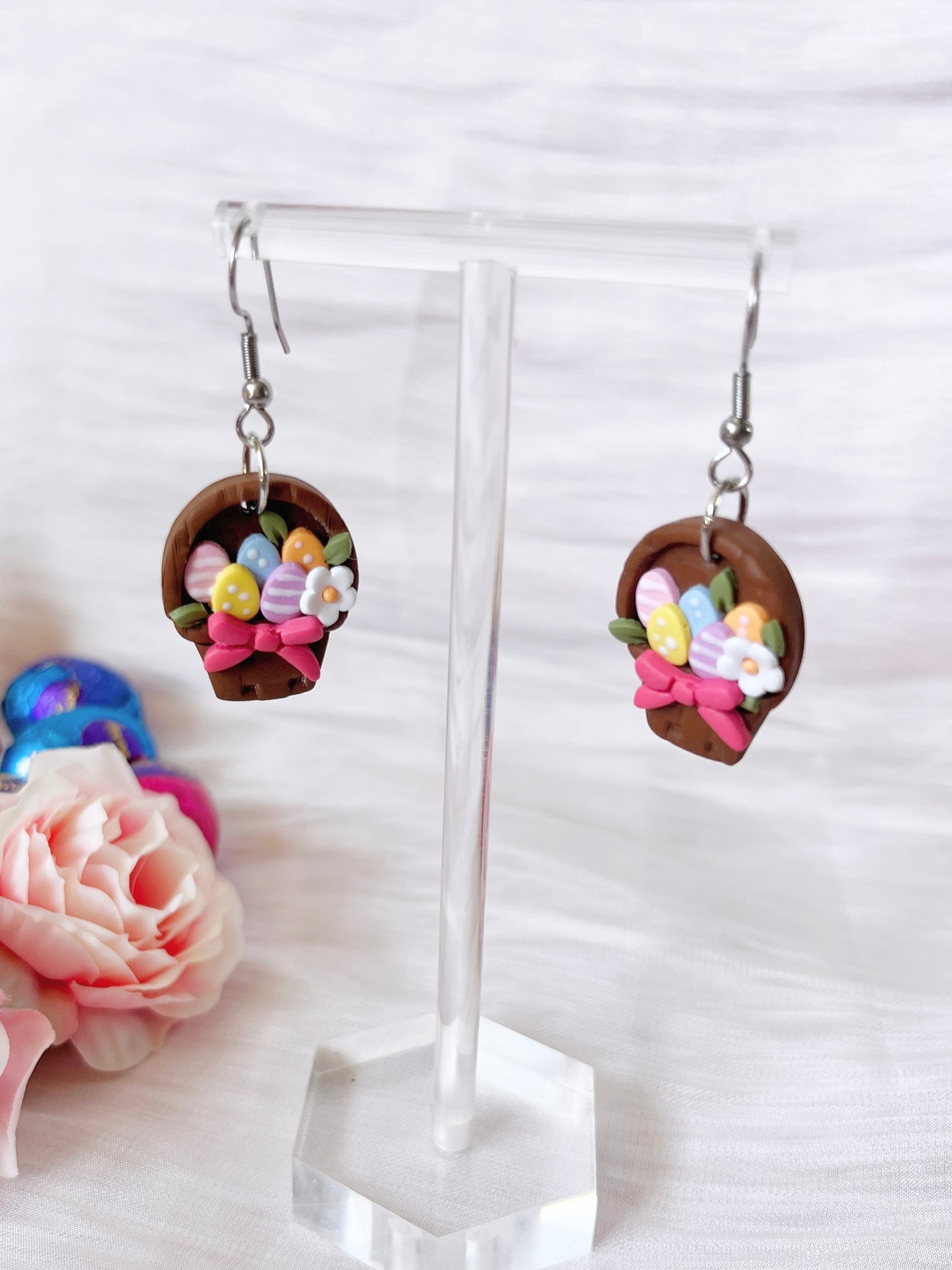 The Egg Basket Polymer Clay Earrings by silverstar.nz are handmade and feature basket shapes filled with colorful eggs and flowers. Displayed on a clear stand, they are set against a background of a pink rose and soft white fabric, highlighting their unique design.