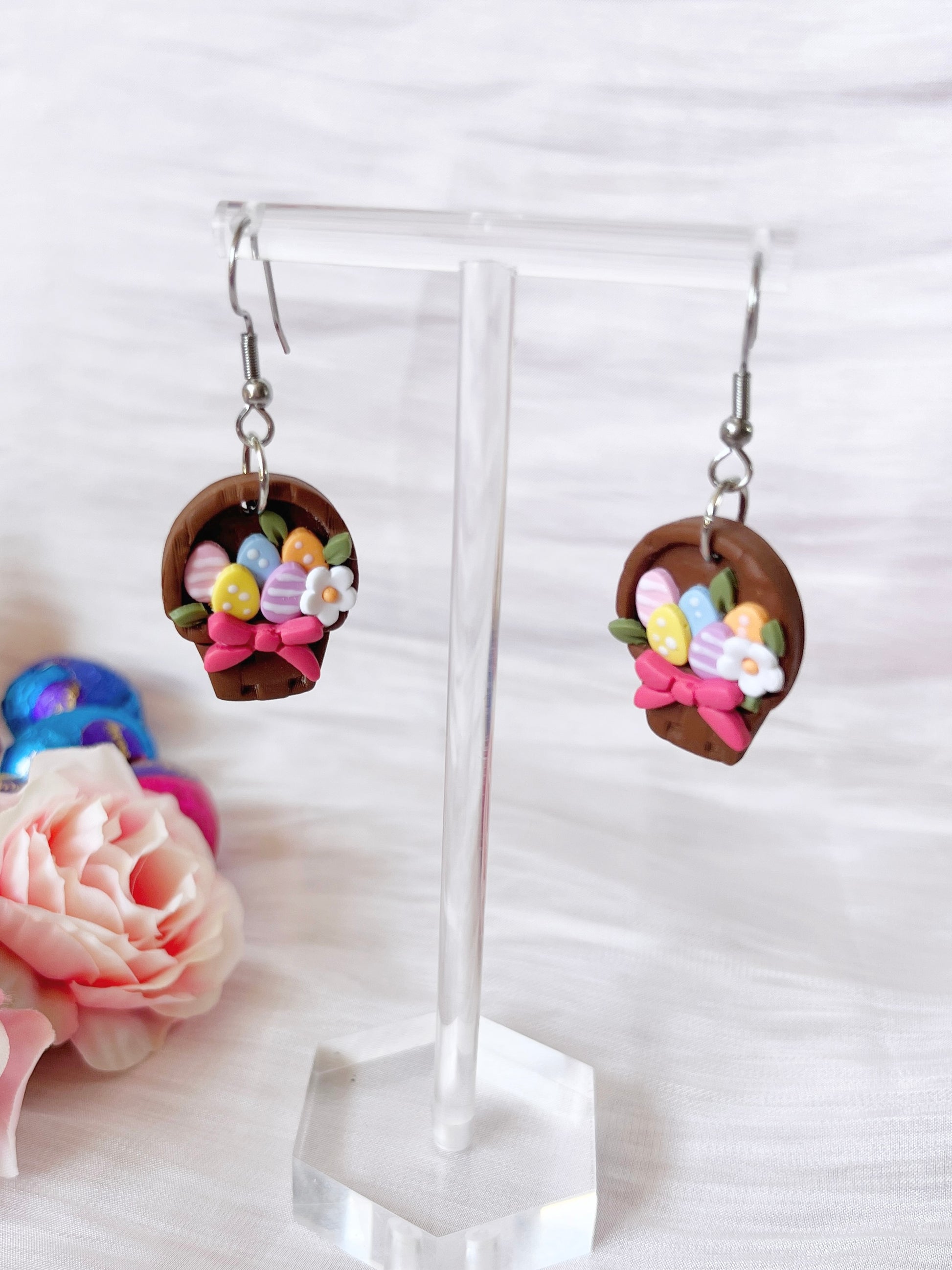 The Egg Basket Polymer Clay Earrings by silverstar.nz are handmade and feature basket shapes filled with colorful eggs and flowers. Displayed on a clear stand, they are set against a background of a pink rose and soft white fabric, highlighting their unique design.