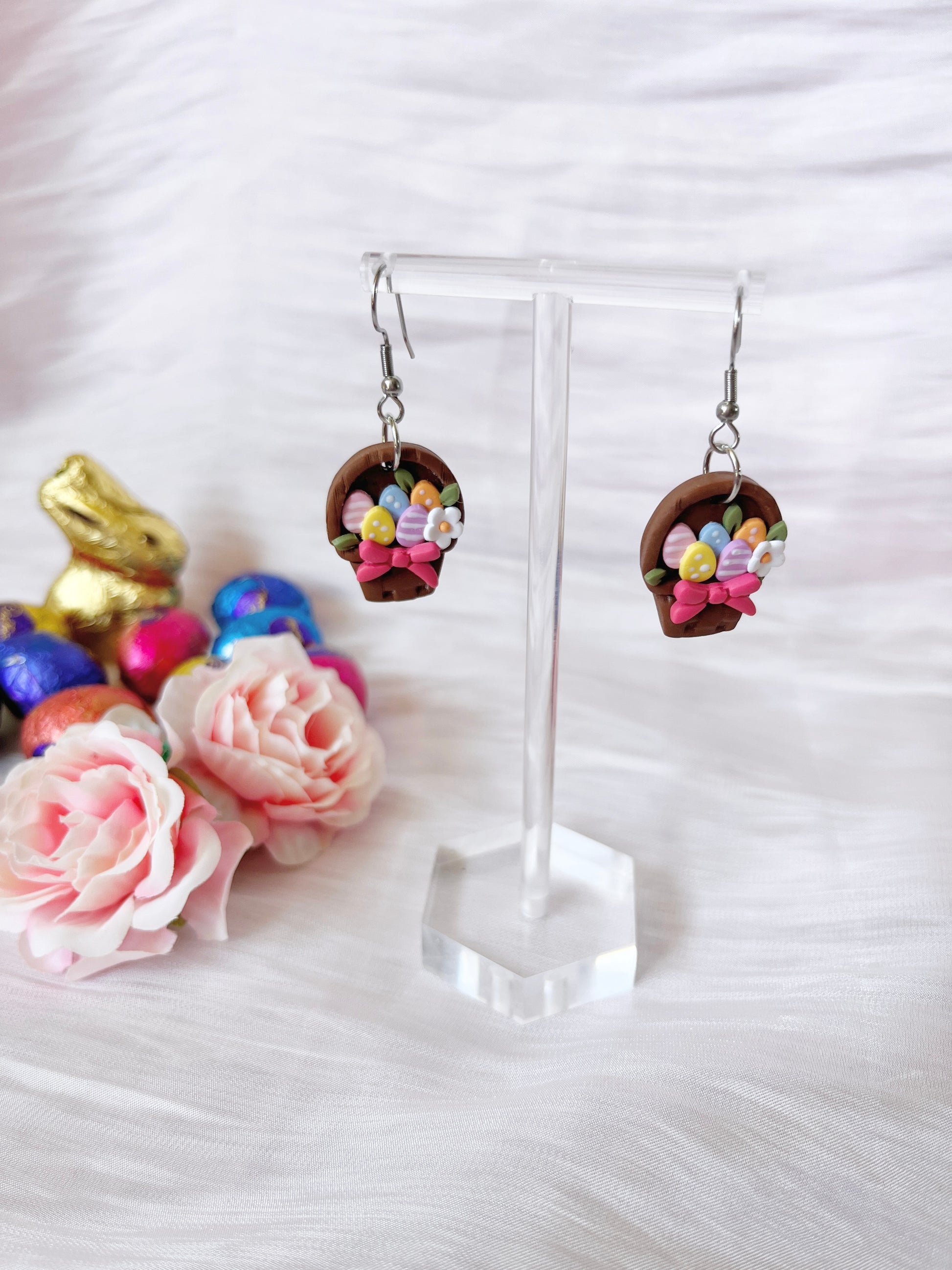 The Egg Basket Polymer Clay Earrings by silverstar.nz are displayed on a clear stand, surrounded by pink roses and Easter-themed decor like a gold bunny and colorful eggs, all set on white textured fabric. These earrings are handmade and shaped like small baskets filled with colorful eggs.