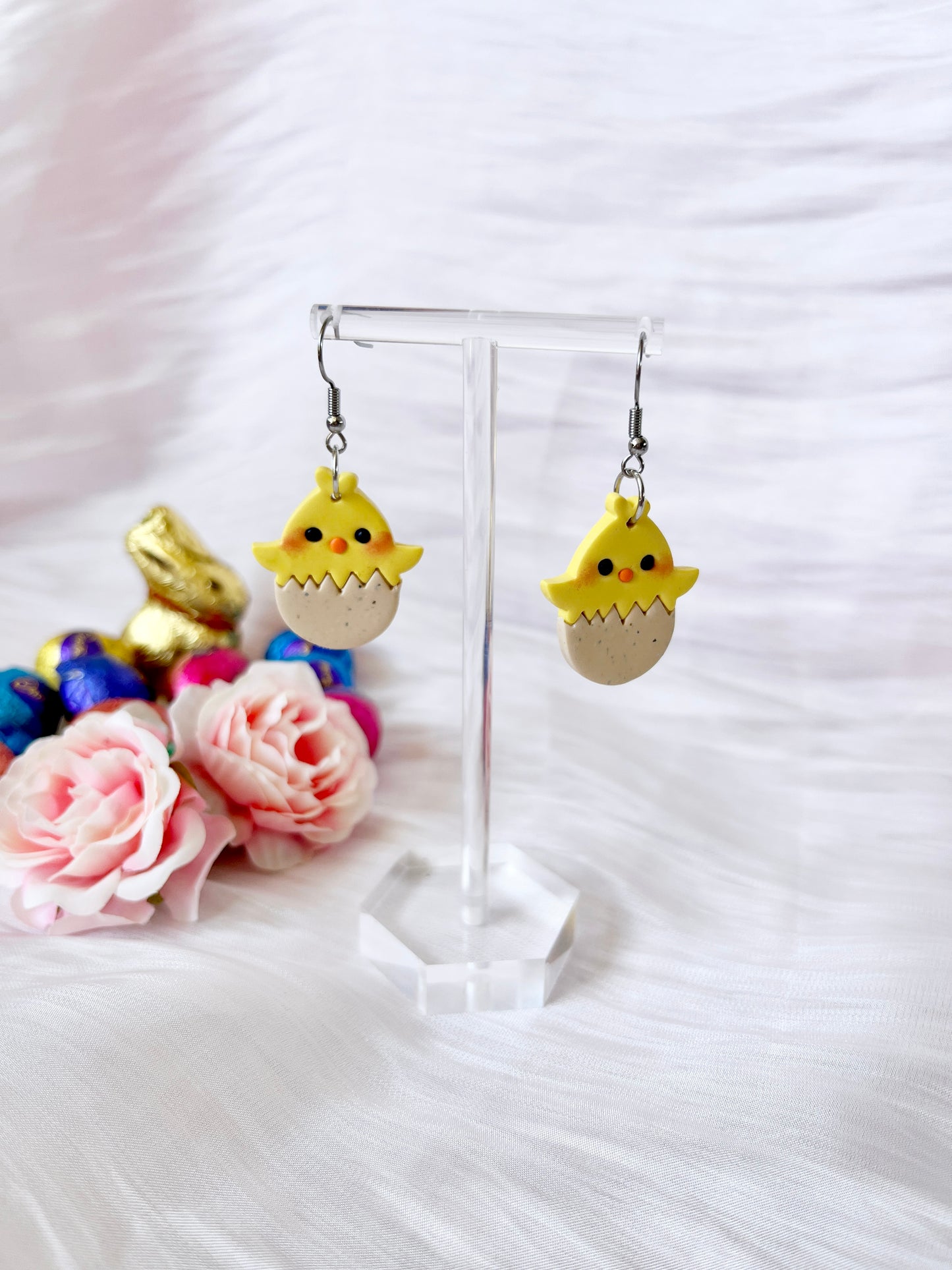 The Egg Baby Chick Polymer Clay Earrings by silverstar.nz, featuring a unique design of yellow chicks hatching from eggs, are displayed on a stand. Beside them lie pink roses and colorful chocolate eggs, with a small gold chocolate rabbit in the background on a white fabric surface.