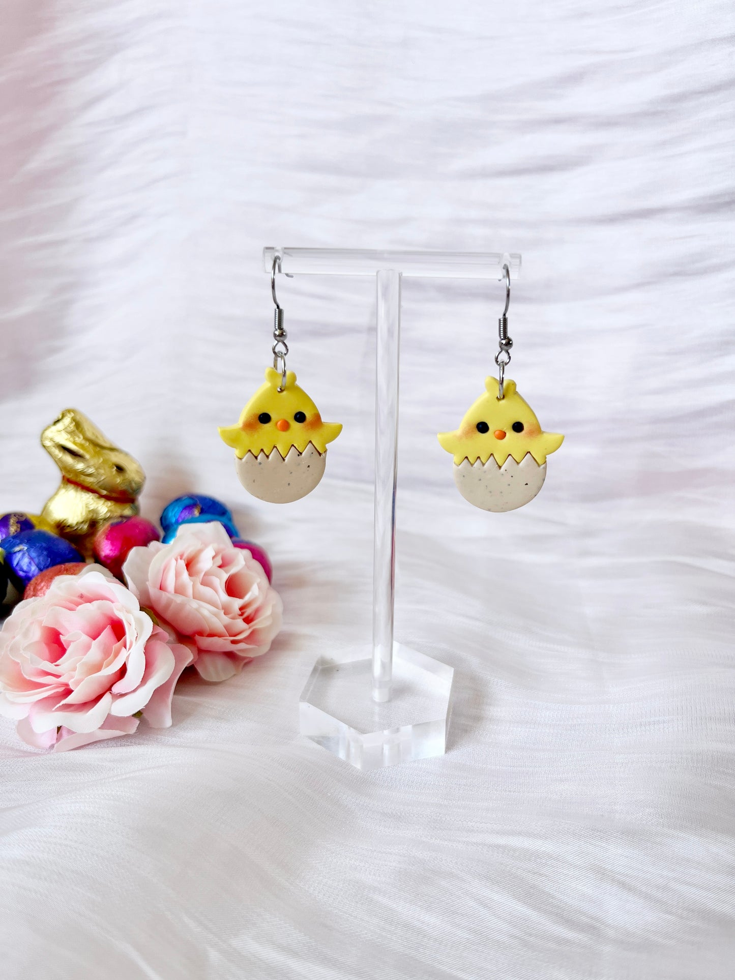Egg Baby Chick Polymer Clay Earrings by silverstar.nz feature yellow chicks hatching from eggs on a clear stand, highlighted by a gold bunny and colorful eggs, with pink roses on a white textured surface in the background.