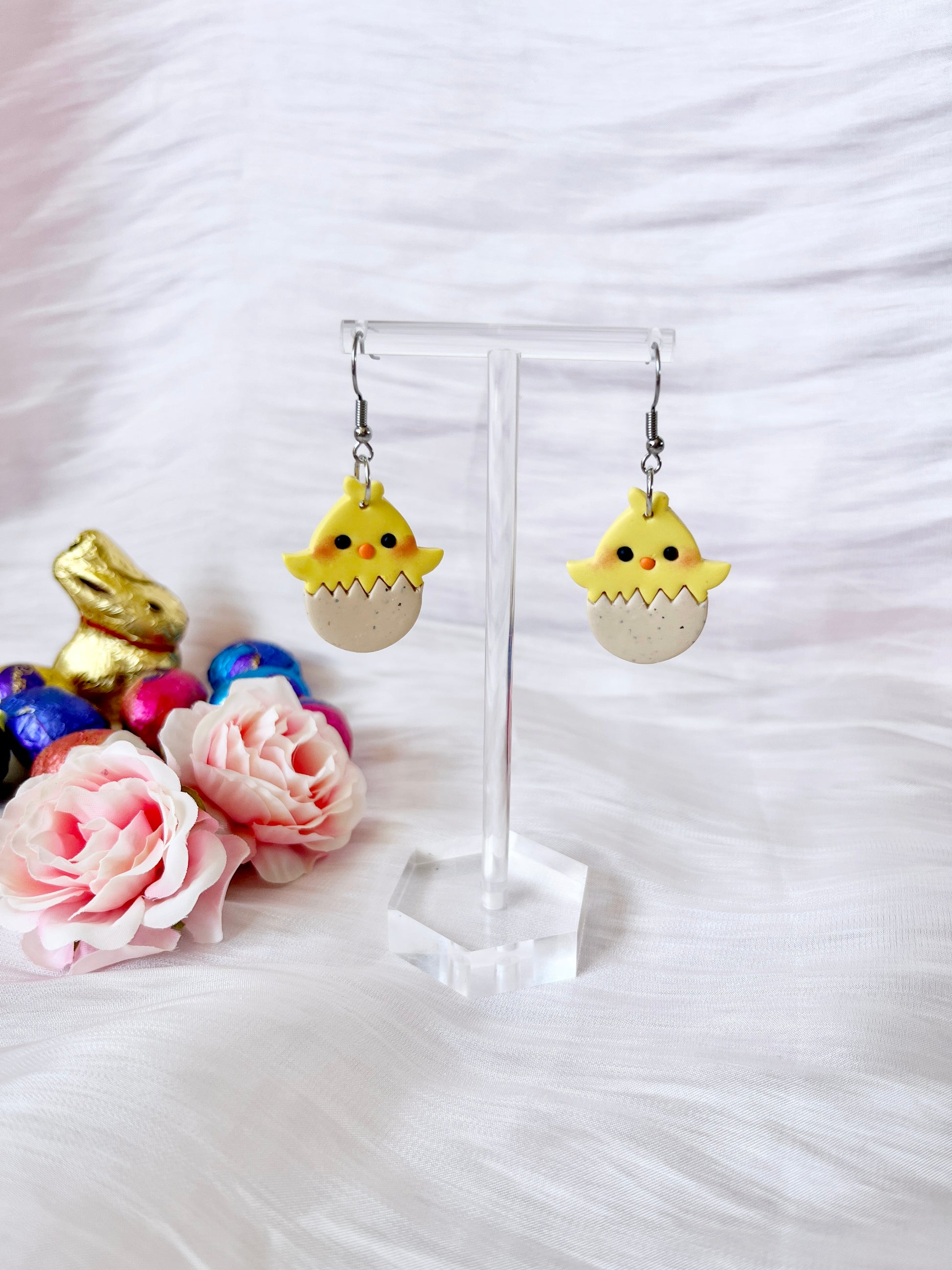 Egg Baby Chick Polymer Clay Earrings by silverstar.nz feature yellow chicks hatching from eggs on a clear stand, highlighted by a gold bunny and colorful eggs, with pink roses on a white textured surface in the background.